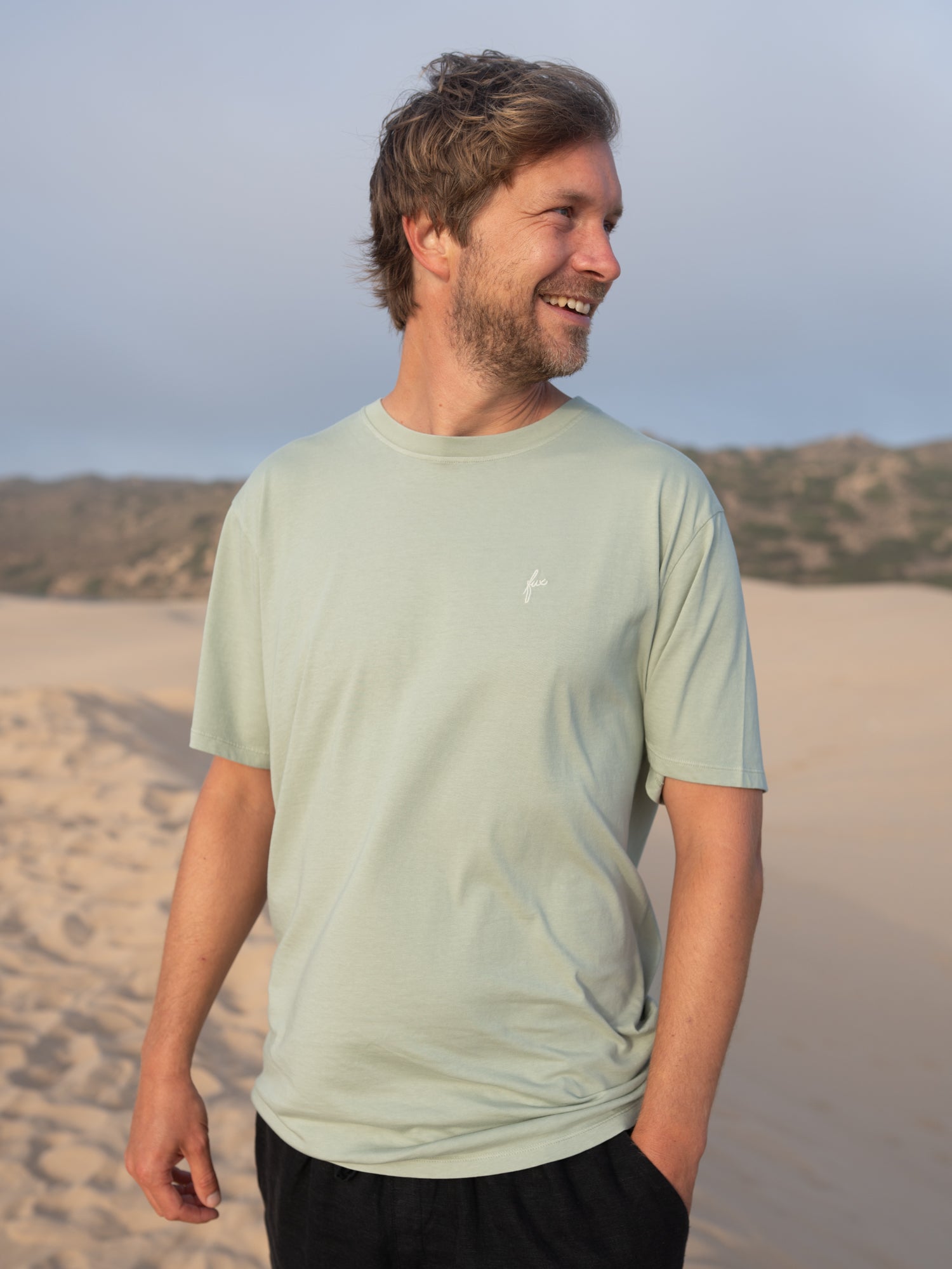 Mann trägt fair gefertigtes seegrünes FUXBAU T-Shirt aus Biobaumwolle vor Dünenlandschaft