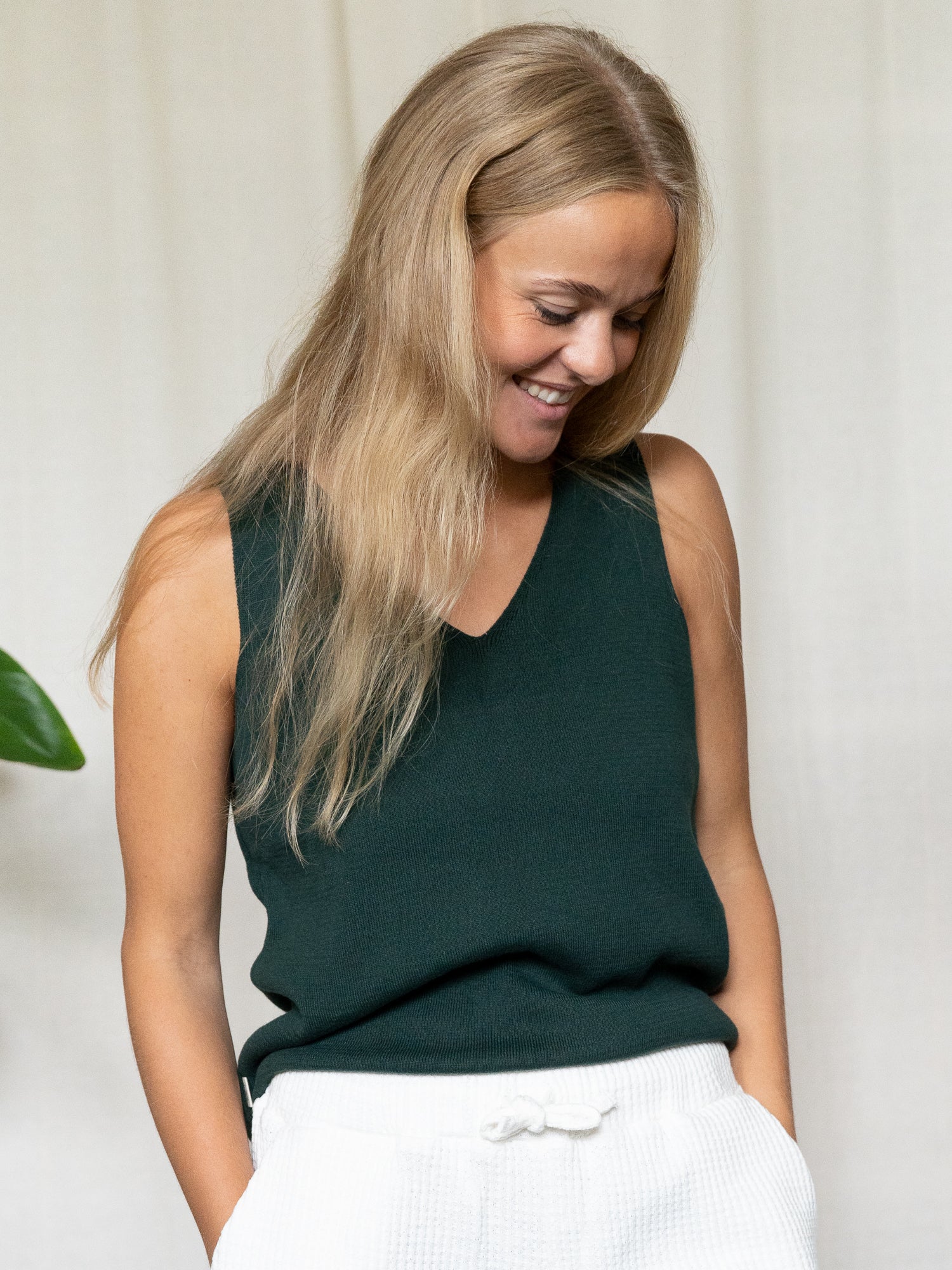 Frau mir dunkelgrünem Stricktop und weißer Shorts hat die Händen in den Taschen und lächelt mit dem Blick nach unten gerichtet.