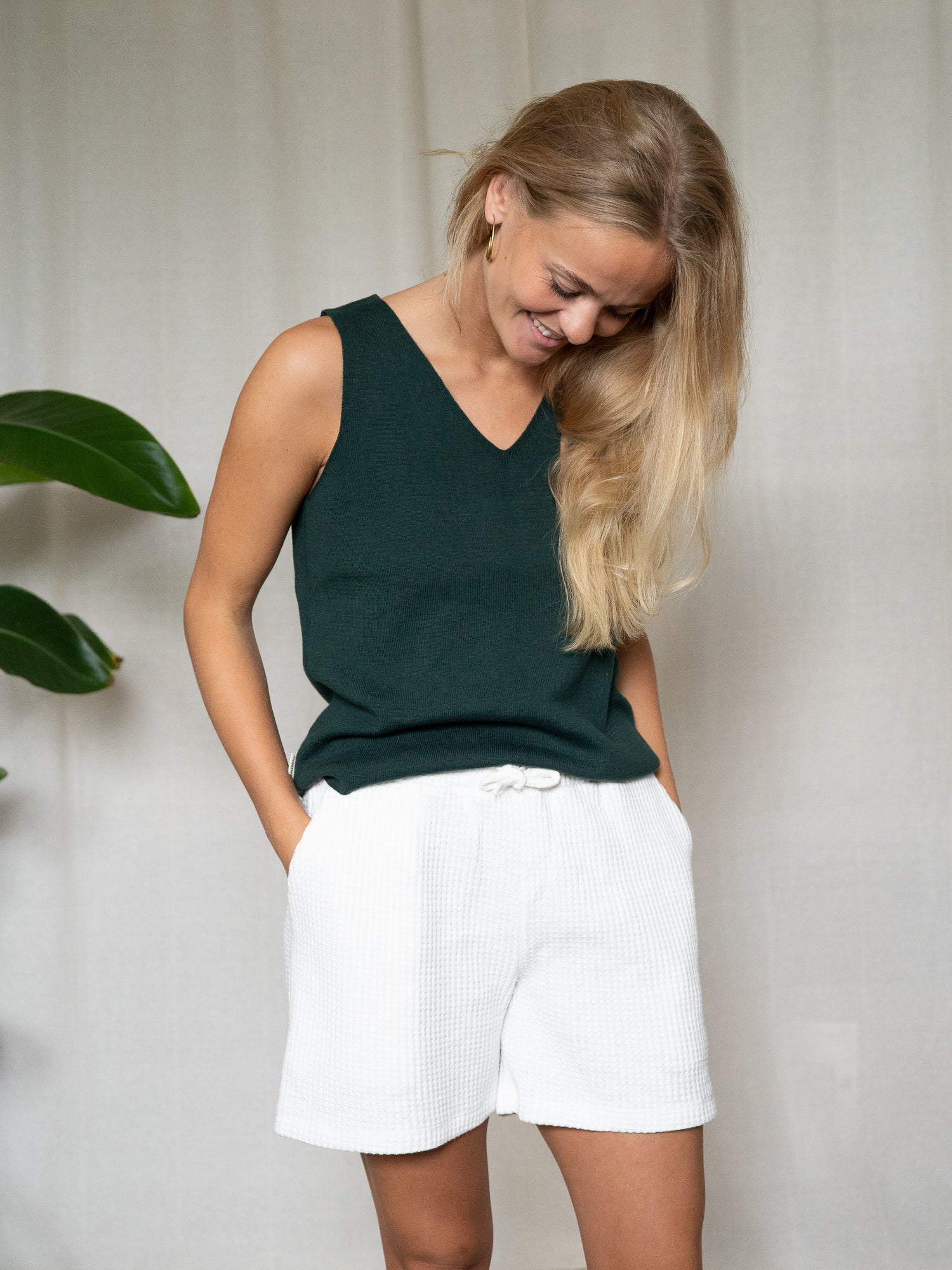 Frau mit dunkelgrünem Stricktop. welches einen V-Ausschnitt hat, lächelt und schaut nach unten. Sie trägt eine weiße Waffel strick Shorts und hat die Hände in den Taschen.