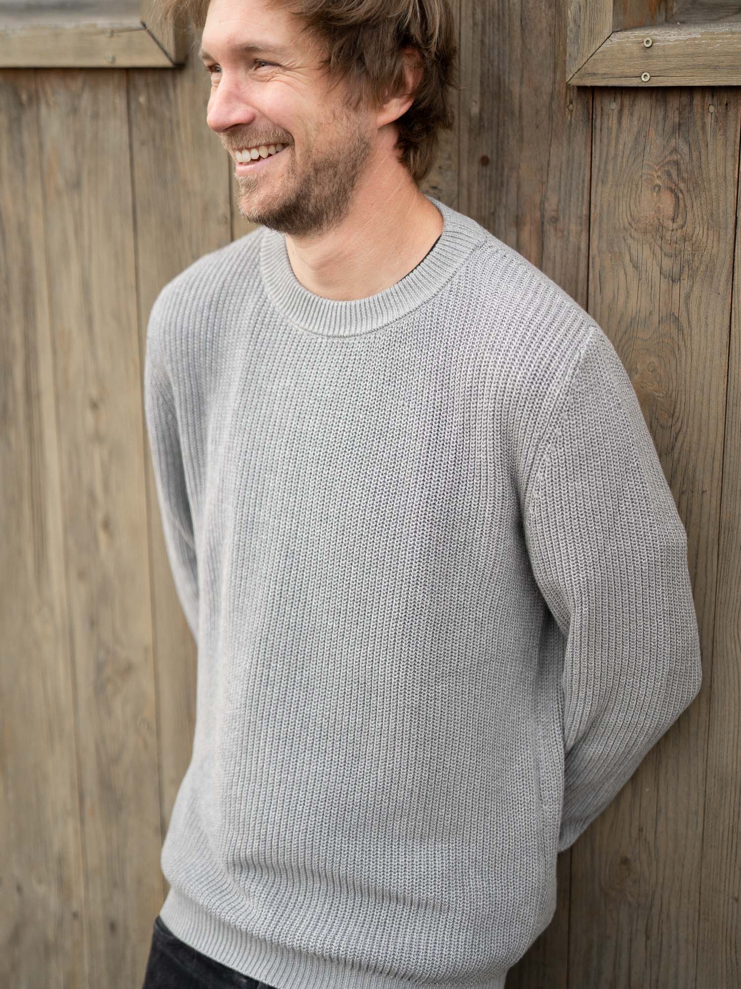 Junger, blonder Mann trägt einen nachhaltig und fair gefertigen 4Seasons Strickpullover von FUXBAU aus 100% GOTS zertifizierte Biobaumwolle in grau.
