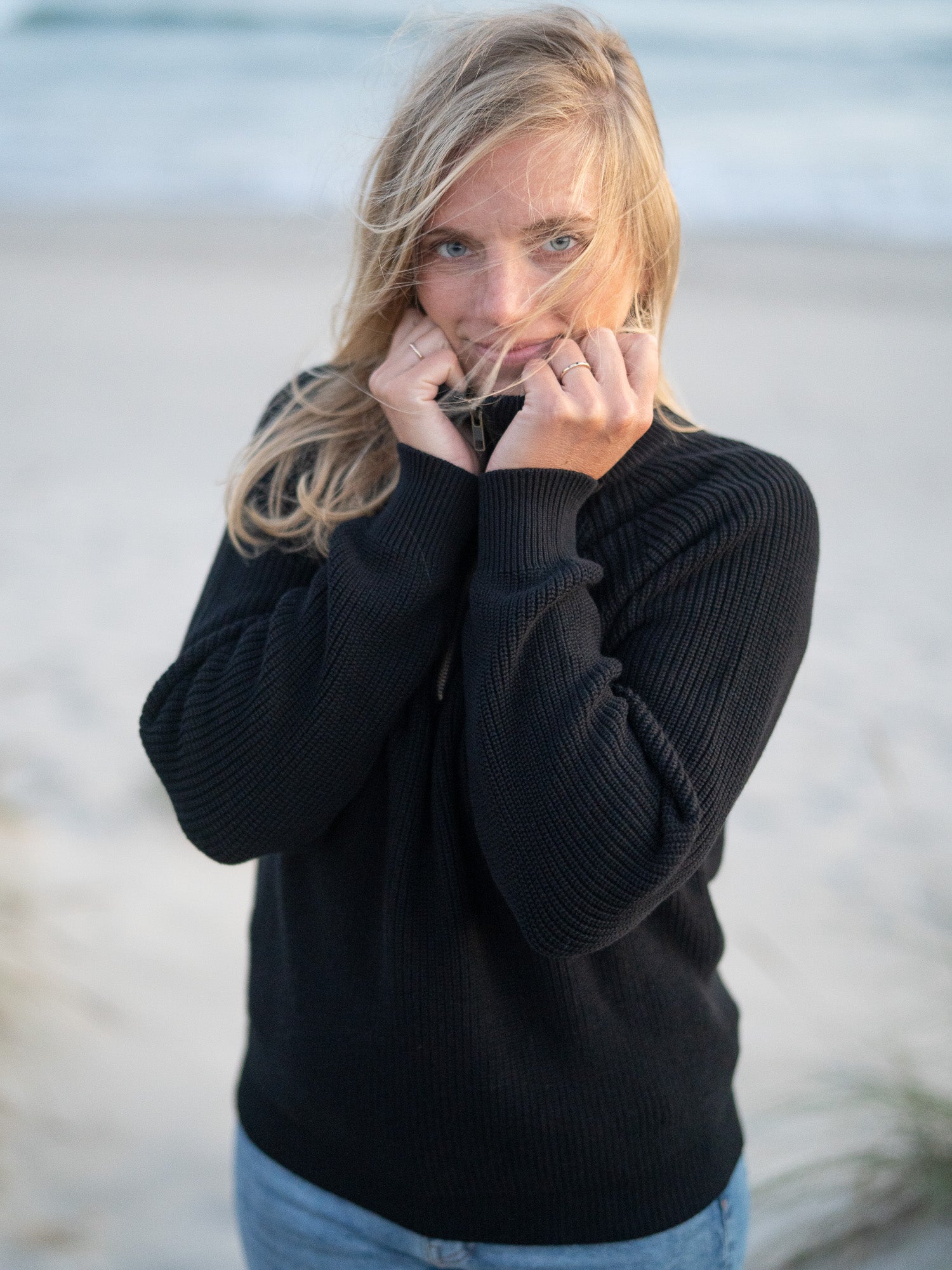 Eine junge, blonde Frau trägt einen nachhaltigen Fair Fashion 4Seasons Troyer aus Biobaumwolle Made in Italy vom nachhaltigen Fair Fashion Label FUXBAU am Strand in Portugal. Frontalansicht.