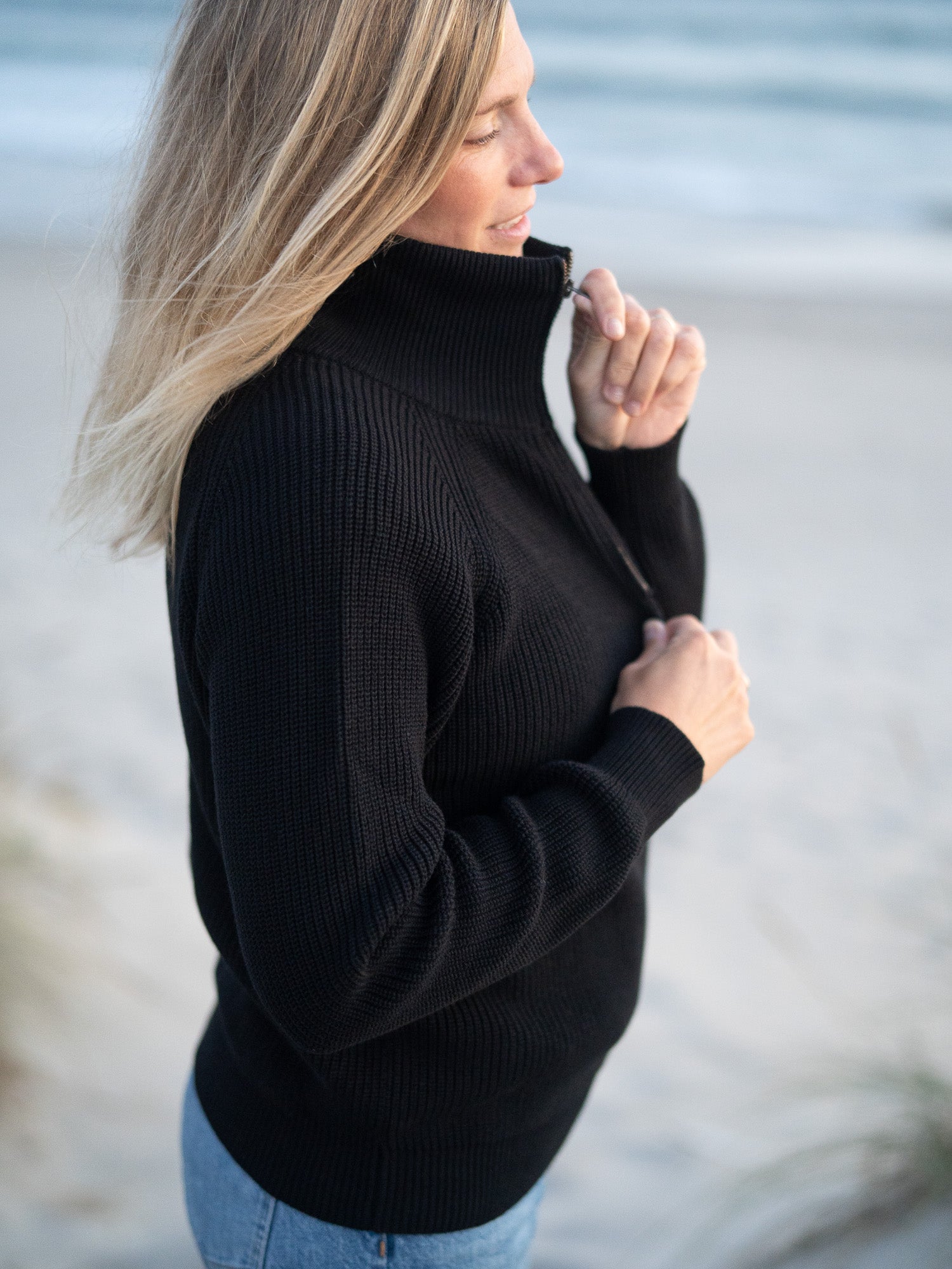 Eine junge, blonde Frau trägt einen nachhaltigen Fair Fashion 4Seasons Troyer aus Biobaumwolle Made in Italy vom nachhaltigen Fair Fashion Label FUXBAU am Strand in Portugal. Seitliche Ansicht.