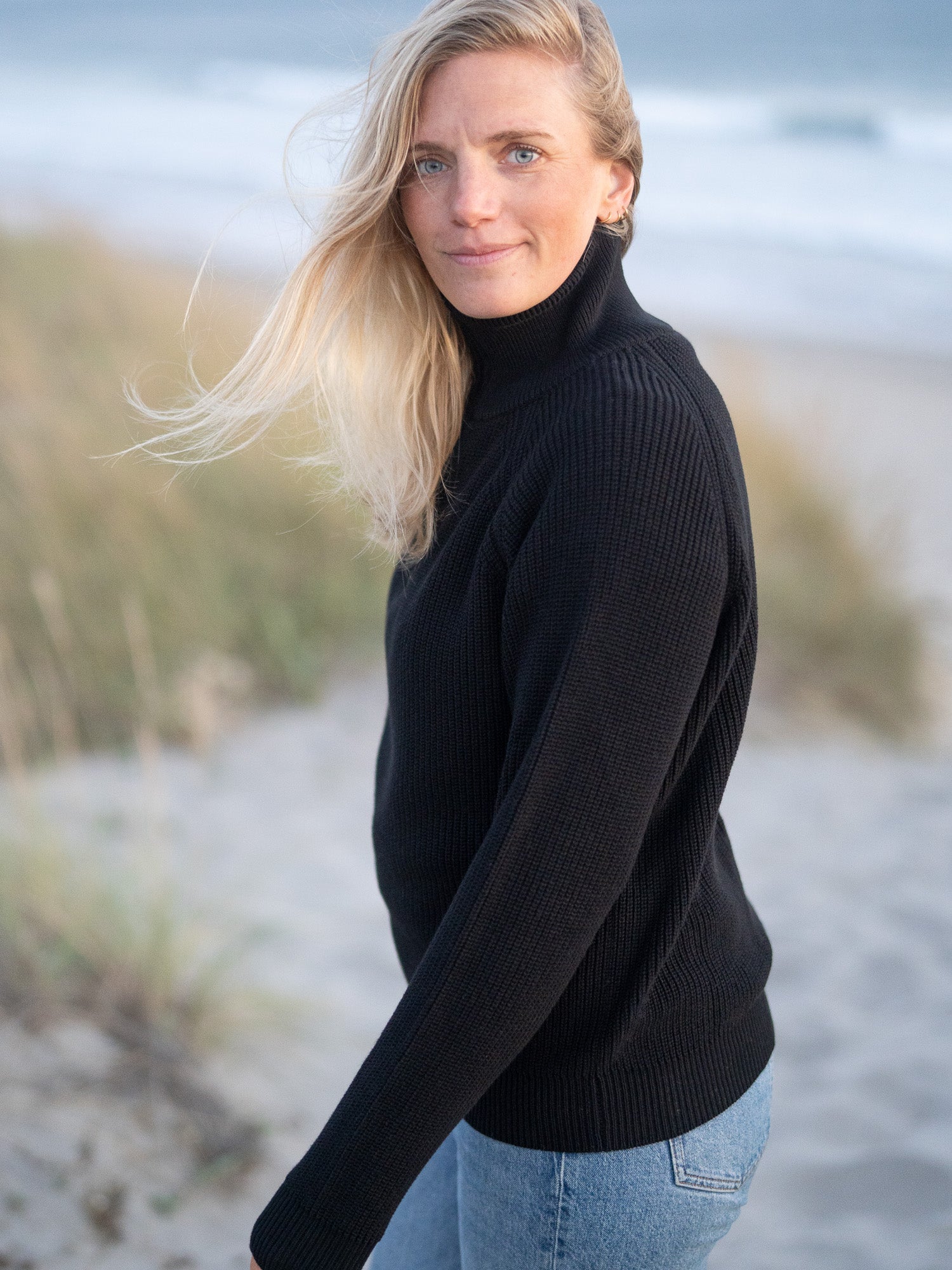 Eine junge, blonde Frau trägt einen nachhaltigen Fair Fashion 4Seasons Troyer aus Biobaumwolle Made in Italy vom nachhaltigen Fair Fashion Label FUXBAU am Strand in Portugal. Seitliche Ansicht.