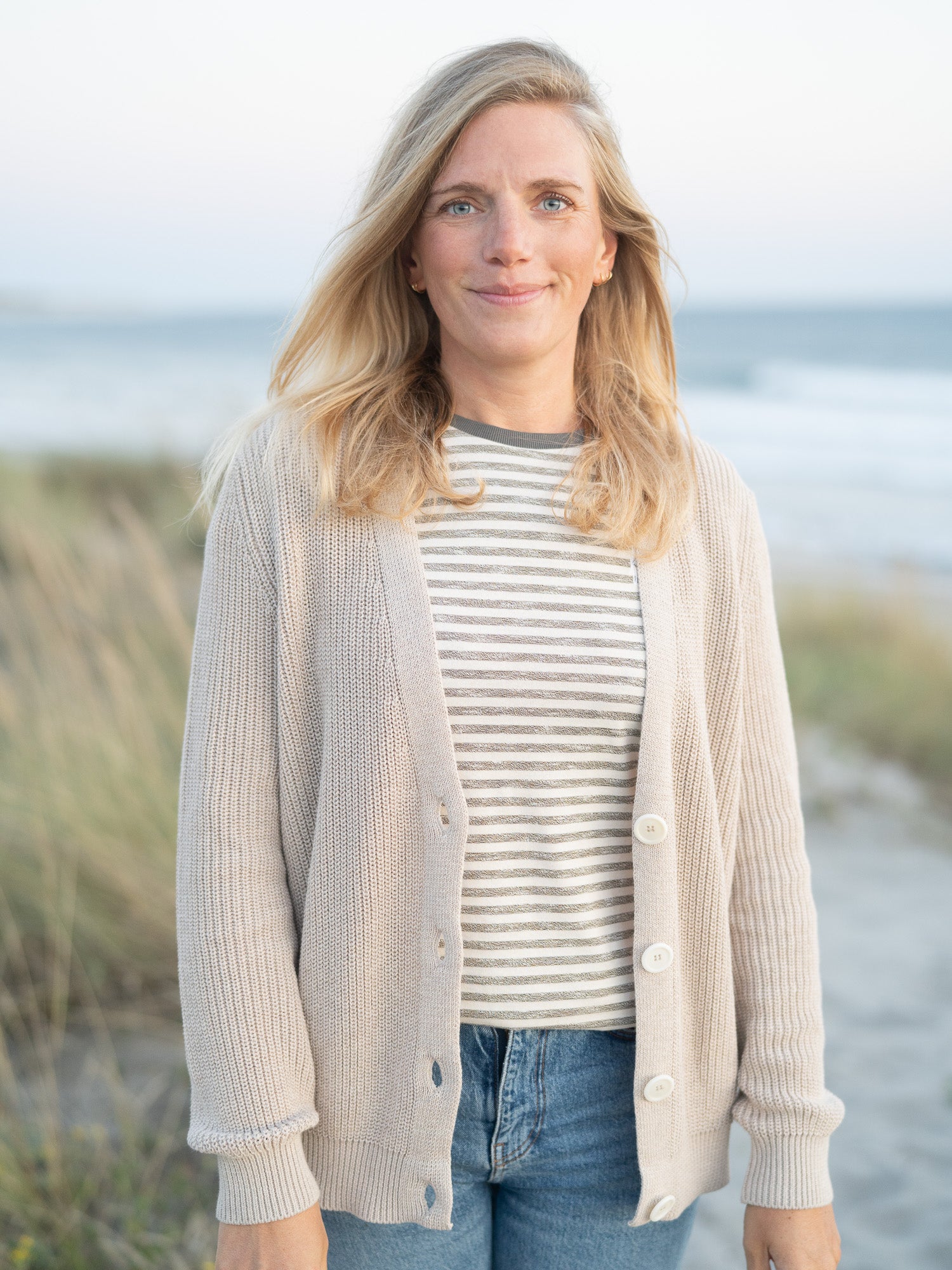 Eine junge, blonde Frau trägt eine beige melierte 4Seasons Strickjacke vom Fair Fashion Modelabel FUXBAU aus 100% GOTS Biobaumwolle am Strand in Portugal.