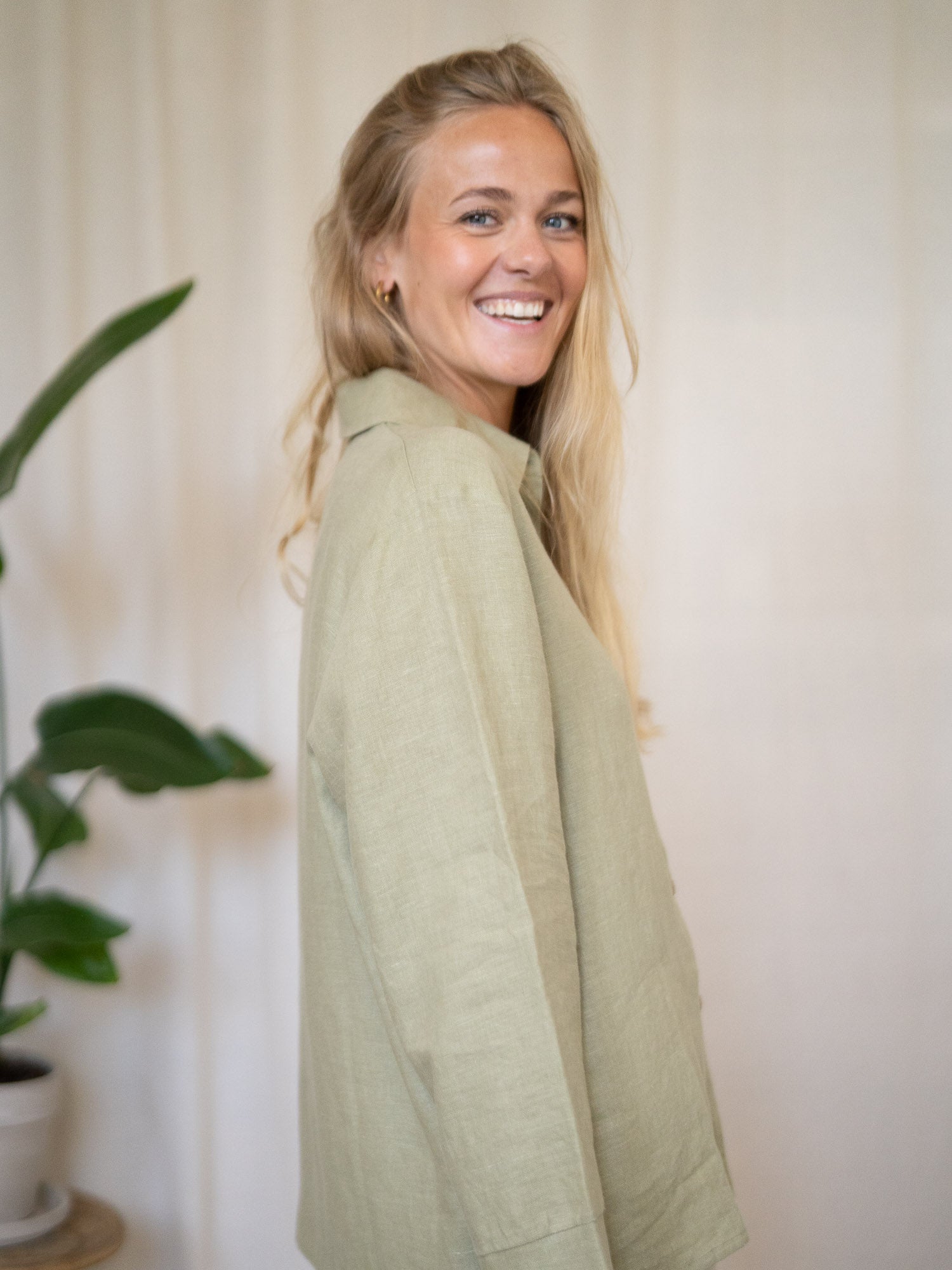 Eine junge, blonde Frau lächelt und steht seitlich zur Kamera. Dabei trägt sie eine nachhaltige Frauen Langarm Leinenbluse aus 100% Leinen. Made in Portugal fair und nachhaltig in der Farbe salbei.