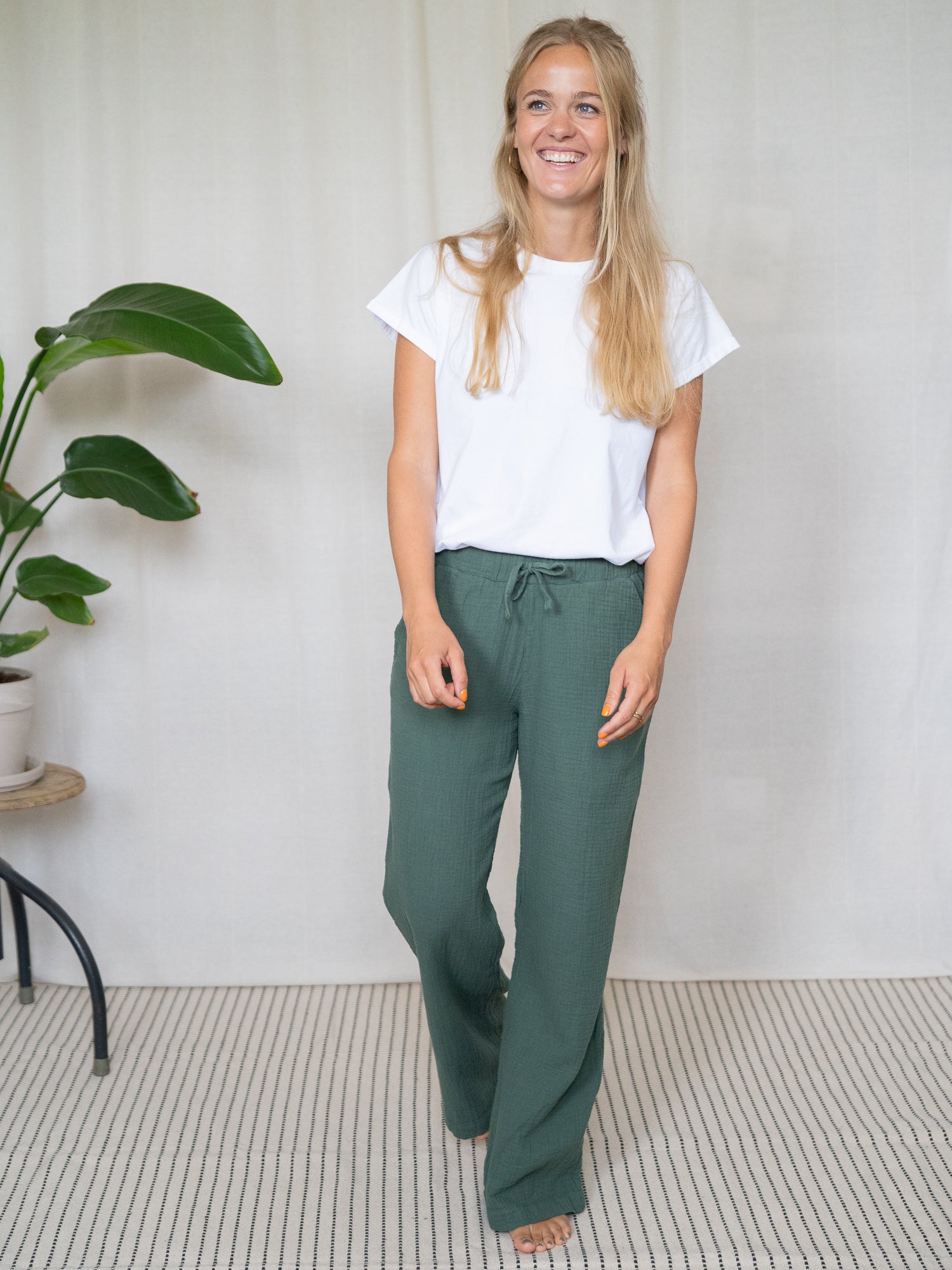 Model mit langer blonder Haaren trägt grüne Musselinhose aus Biobaumwolle und weißes Shirt, stehend vor hellem Vorhang