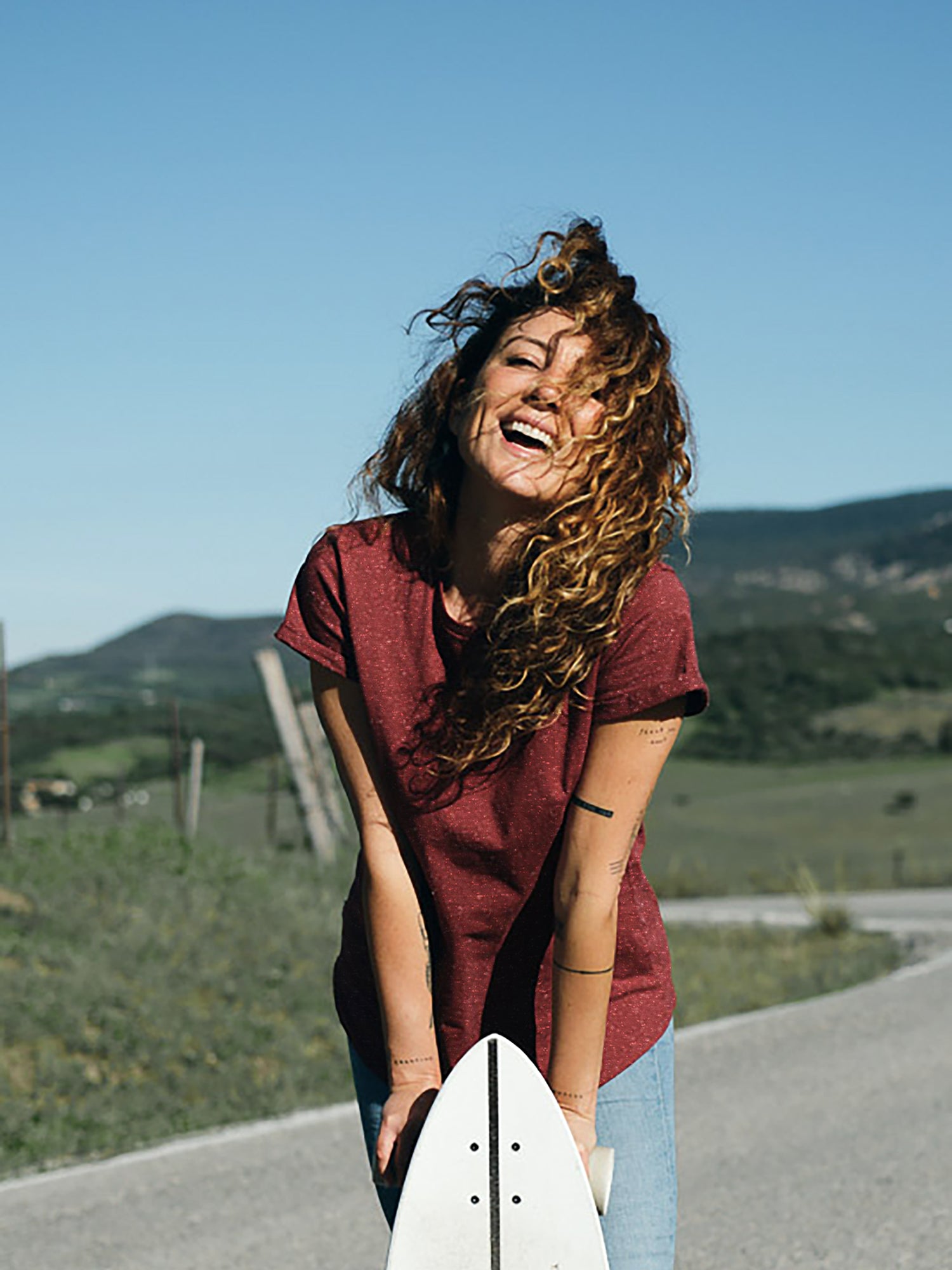 Lockige, braunhaarige Frau mit Longboard in der Hand trägt ein bordeaux farbiges T-Shirt von FUXBAU mit weißen Punkten aus Biobaumwolle.