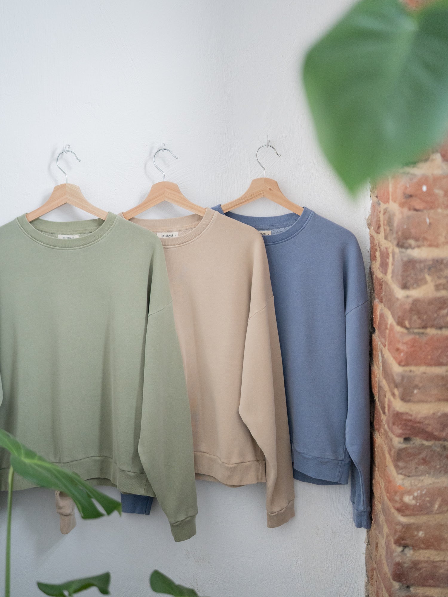 Drei Fair Fashion Frauen oversize Pullover von FUXBAU aus 100% Baumwolle hängen an Kleiderbügeln in den Farben grün, beige, blau an einer weißen Wand.