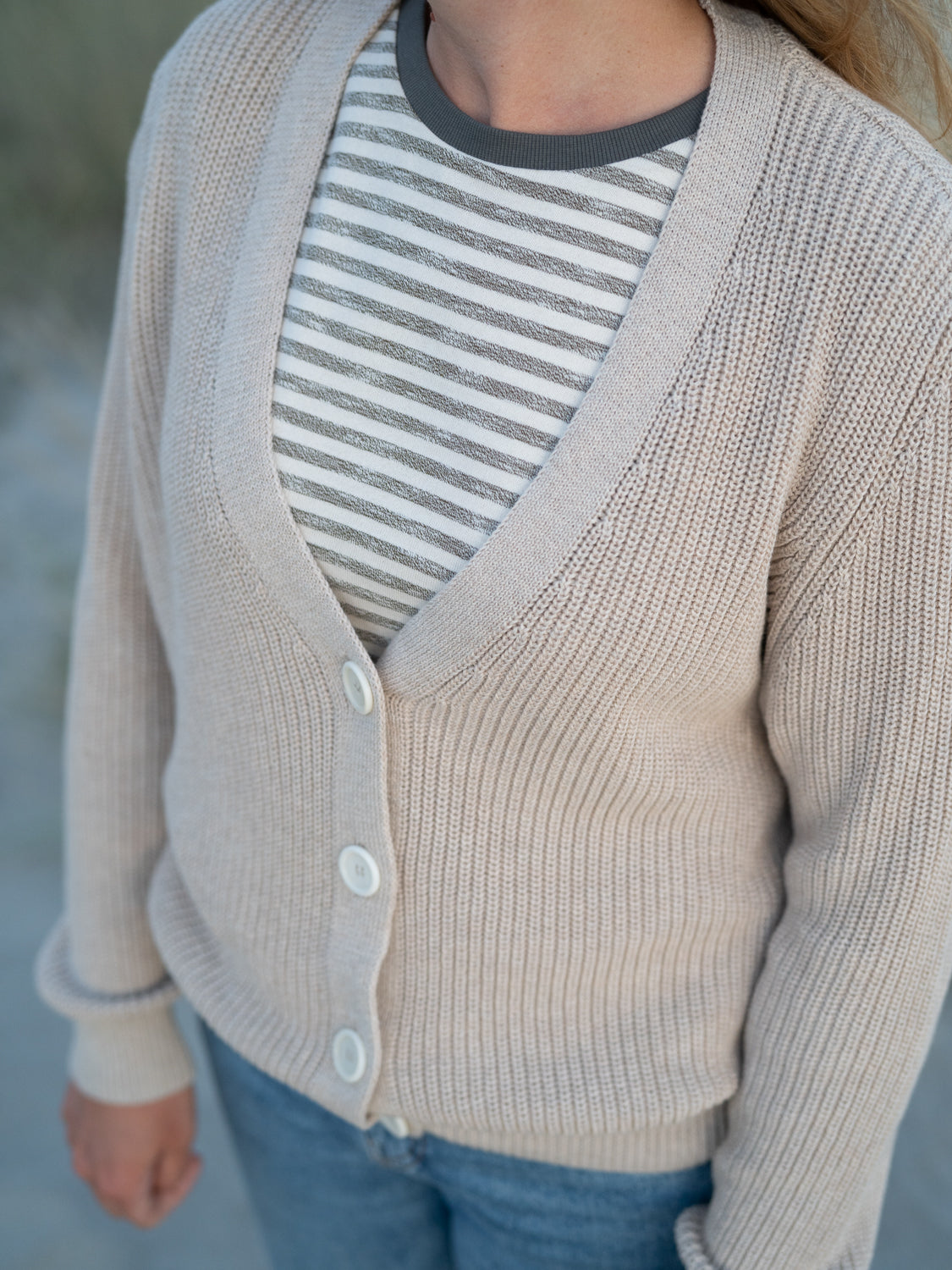 FUXBAU 4Seasons Frauen Strickjacke in beigemeliert aus 100% GOTS zertifizierter Biobaumwolle. Gefertigt in Italien und gertragen von einer jungen Frau am Strand in Portugal.