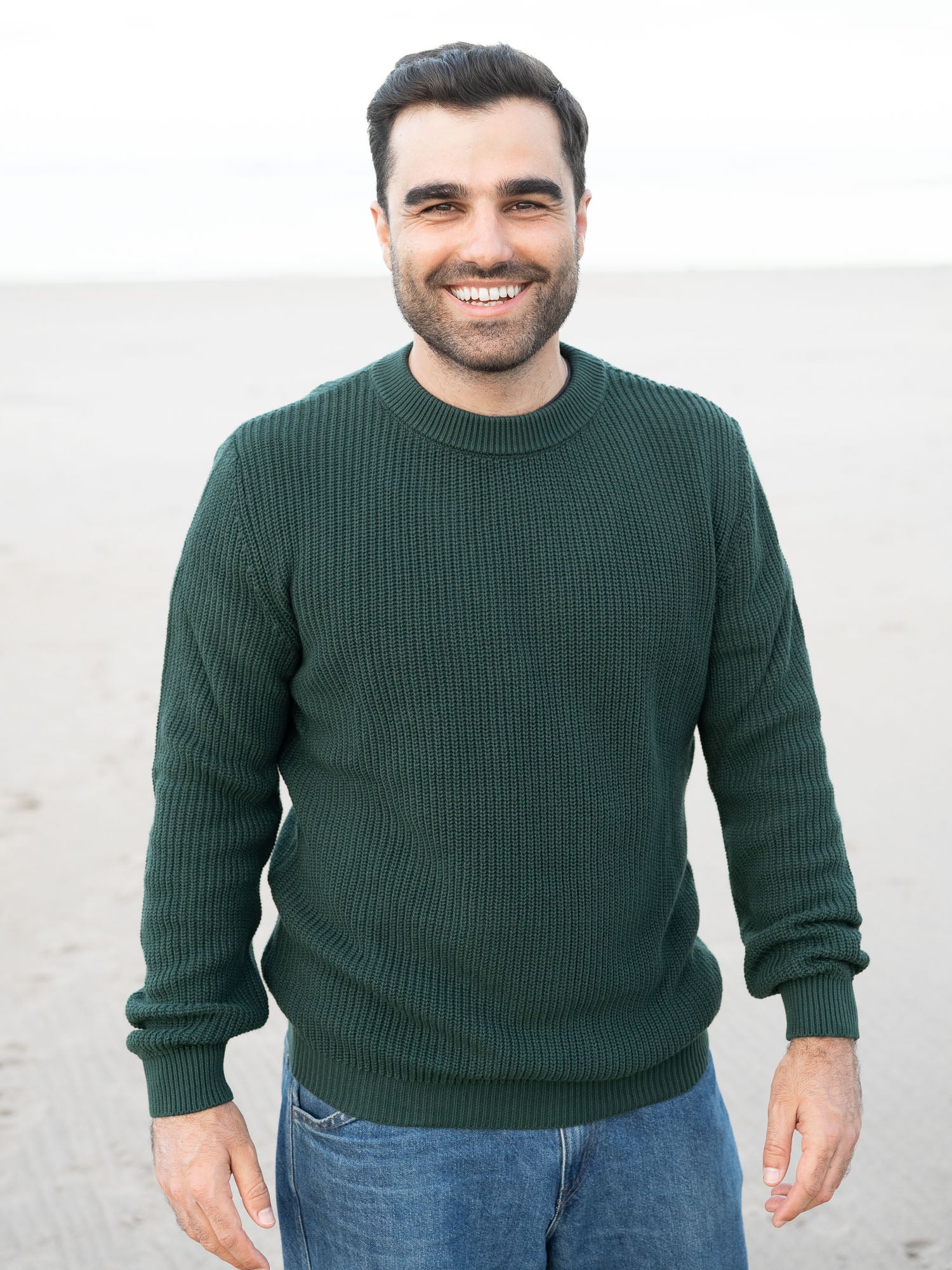 Junger, schwarzhaariger Mann trägt einen nachhaltigen fair fashion Strickpullover von FUXBAU in grün aus GOTS zertifzierter Biobawumwolle am Strand in Spanien.