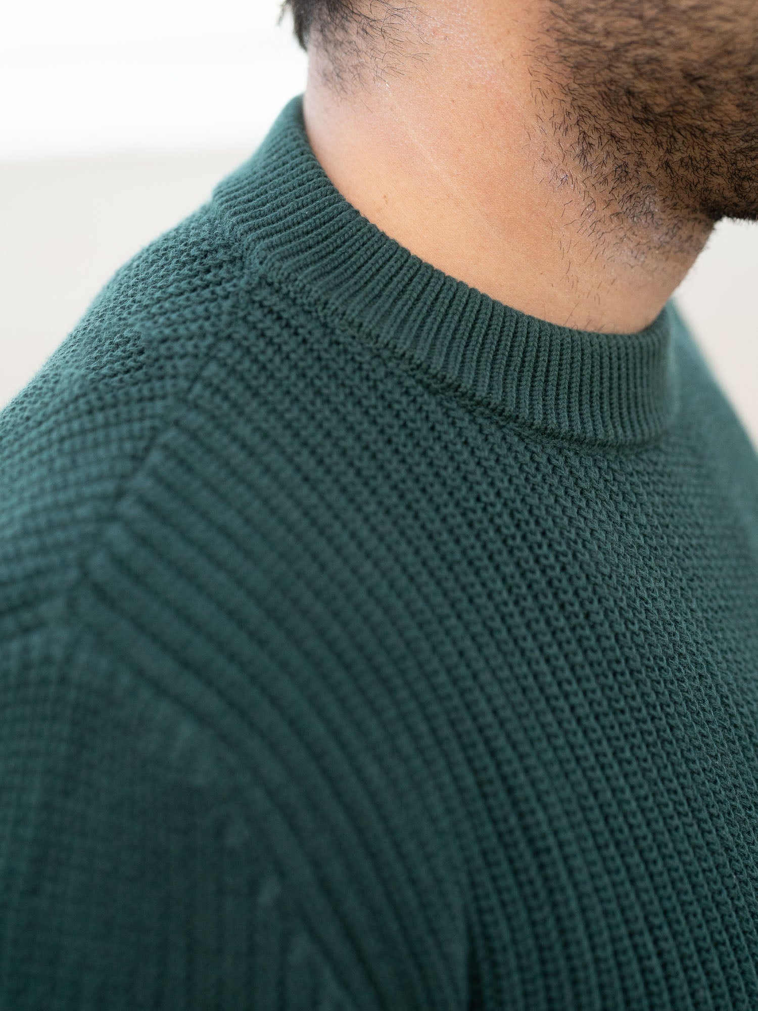 Junger, schwarzhaariger Mann trägt einen nachhaltigen fair fashion Strickpullover von FUXBAU in grün aus GOTS zertifzierter Biobawumwolle am Strand in Spanien.