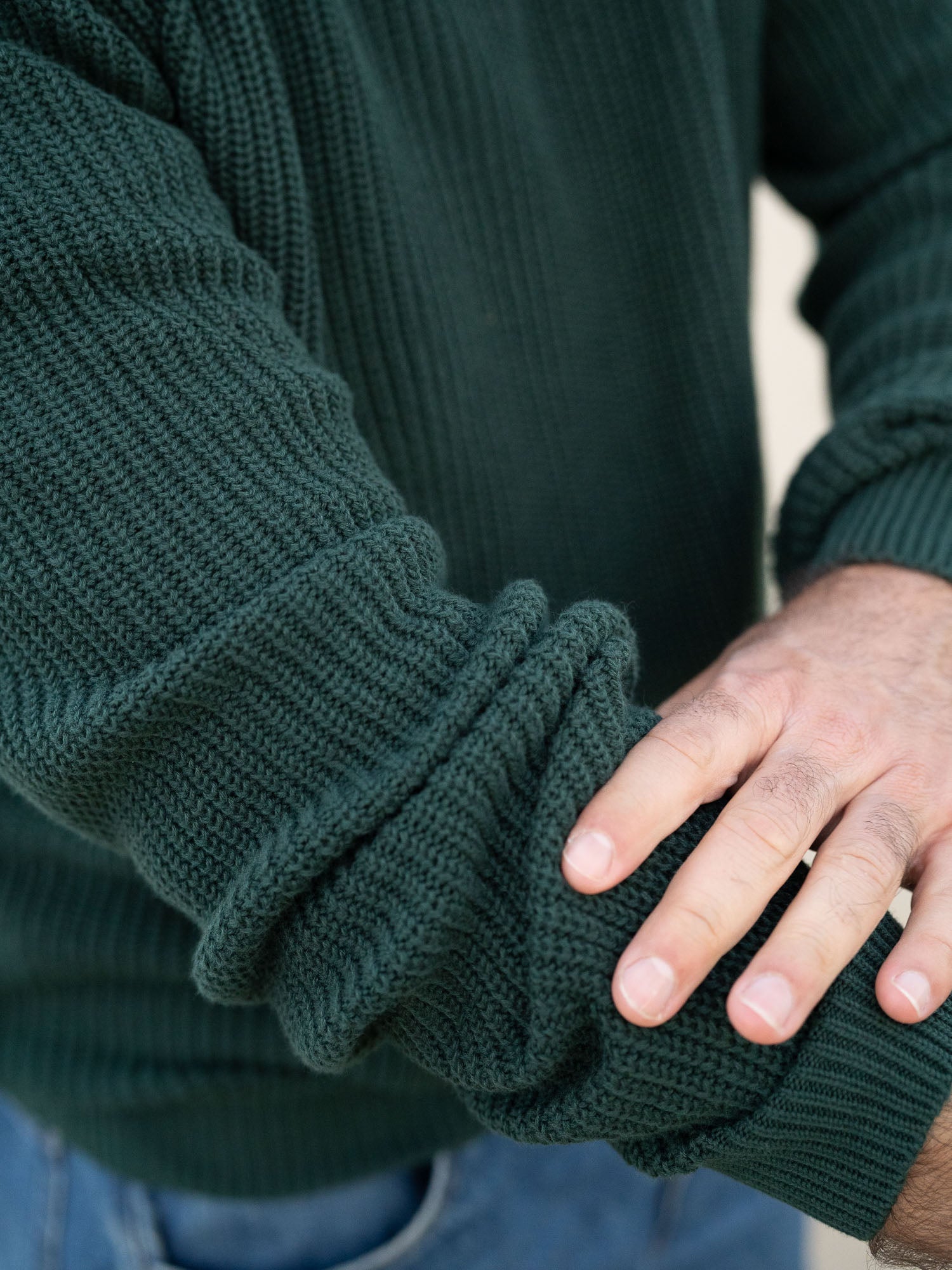 Junger, schwarzhaariger Mann trägt einen nachhaltigen fair fashion Strickpullover von FUXBAU in grün aus GOTS zertifzierter Biobawumwolle am Strand in Spanien.