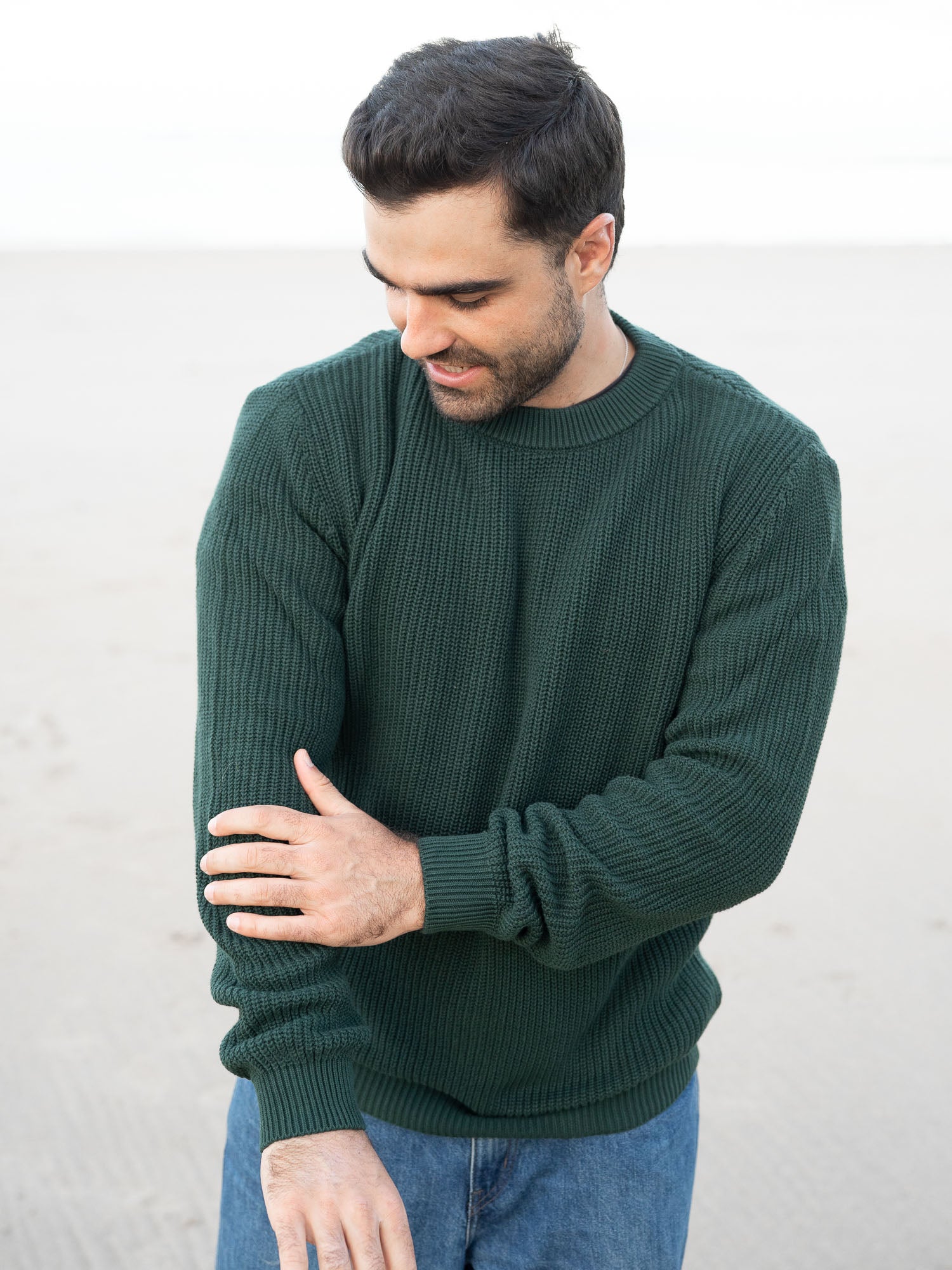 Junger, schwarzhaariger Mann trägt einen nachhaltigen fair fashion Strickpullover von FUXBAU in grün aus GOTS zertifzierter Biobawumwolle am Strand in Spanien.