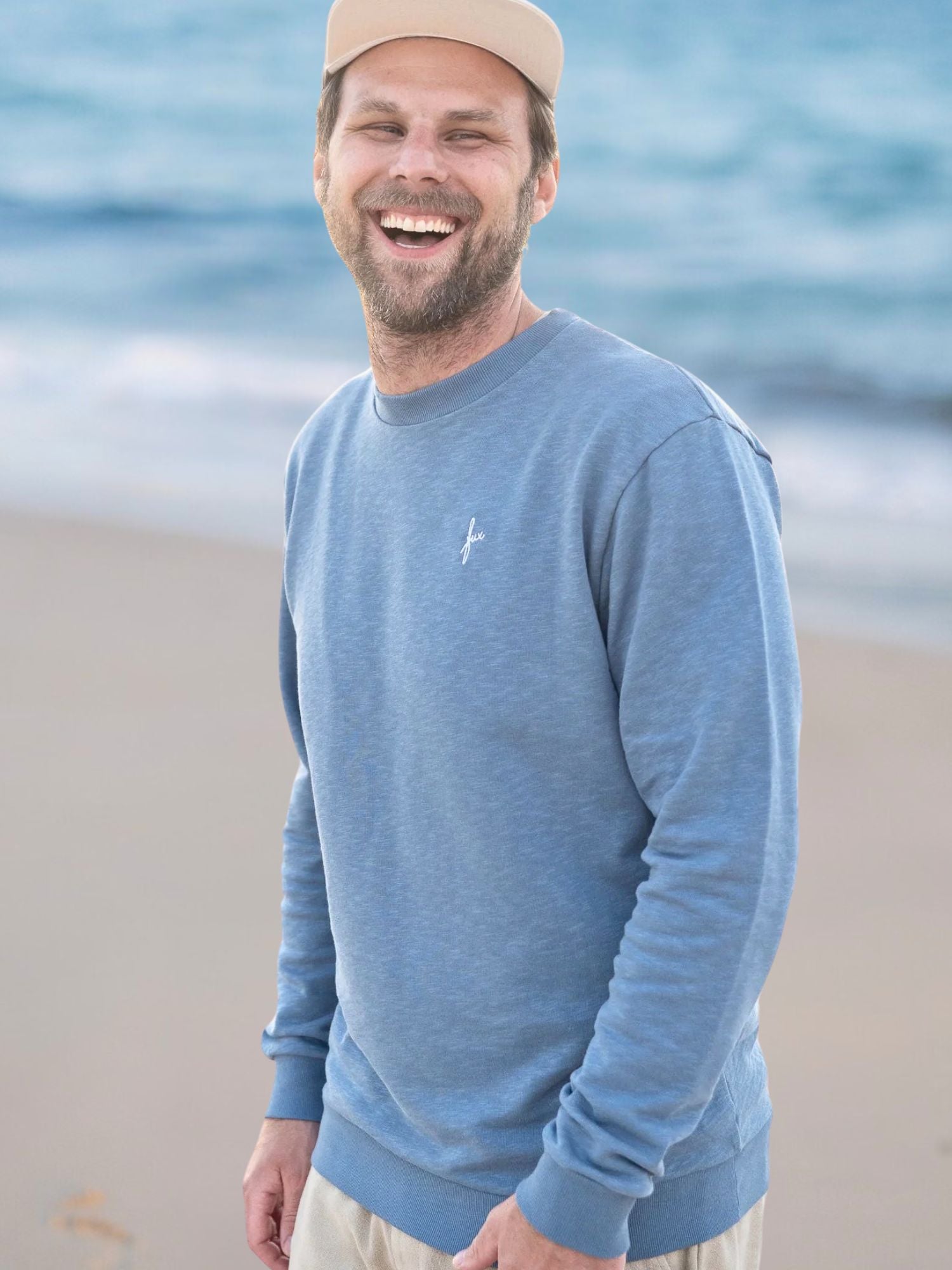 Ein junger Männer trägt einen blau melierten Fair Fashion Pullover am Strand in Portugal aus 100% GOTS zertifizierter Biobaumwolle.