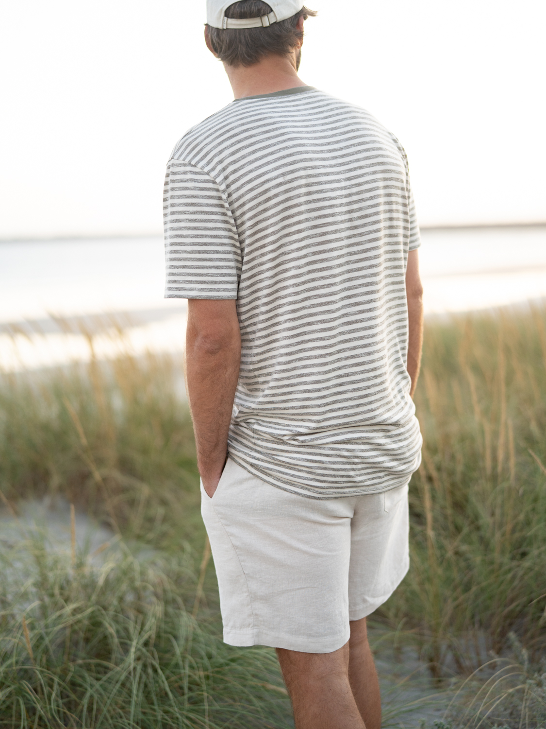 Junger Männ trägt ein nachhaltiges Fair Fashion Outfit von FUXBAU bestehend aus einer creme weissen Leinenshorts und einem grün-beige melierten T-Shirt am Strand in Portugal.