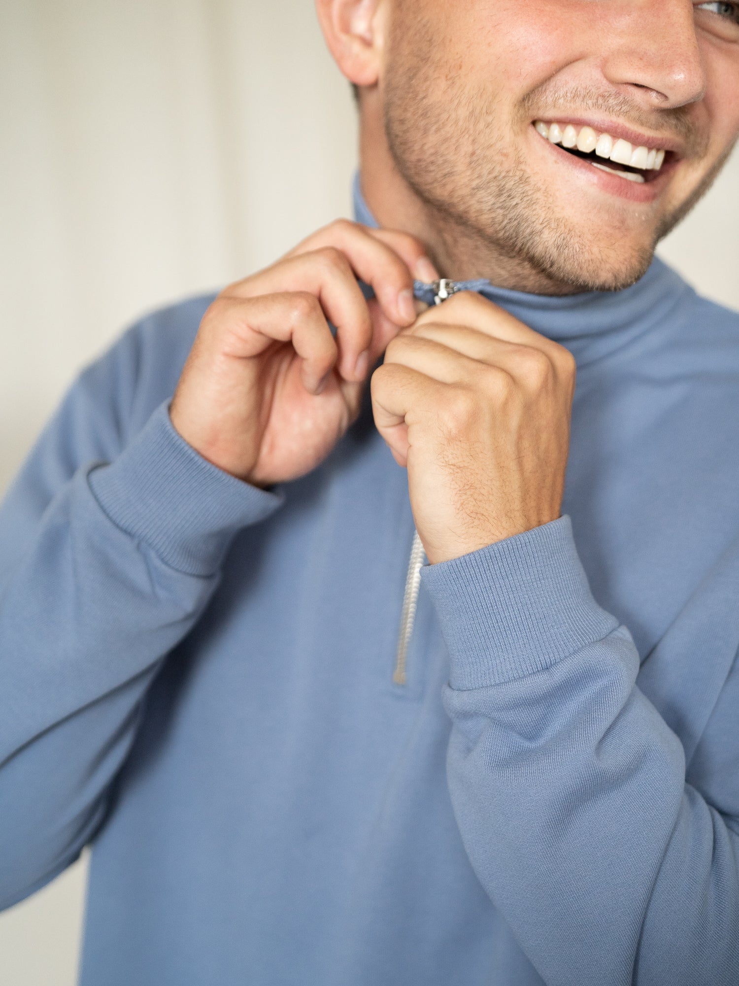 Mann schließt lächelnd den Reißverschluss des himmelblauen Half-Zip Sweaters.