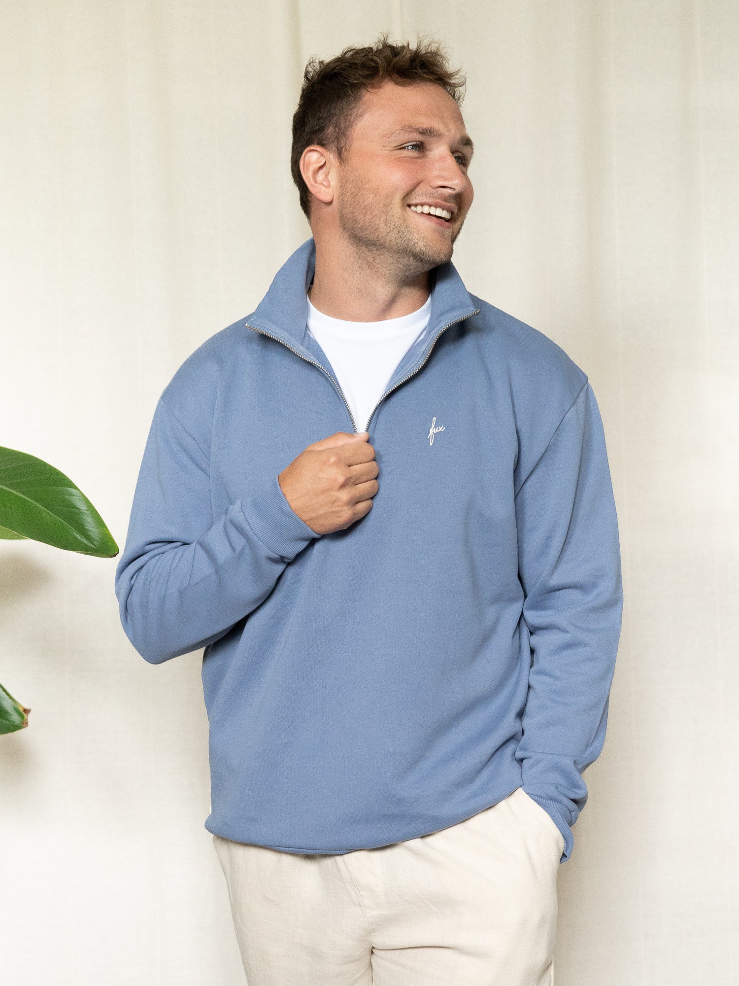 Mann trägt den himmelblauen FUXBAU Half-Zip locker offen über weißem T-Shirt, lächelnd mit Händen in den Taschen.