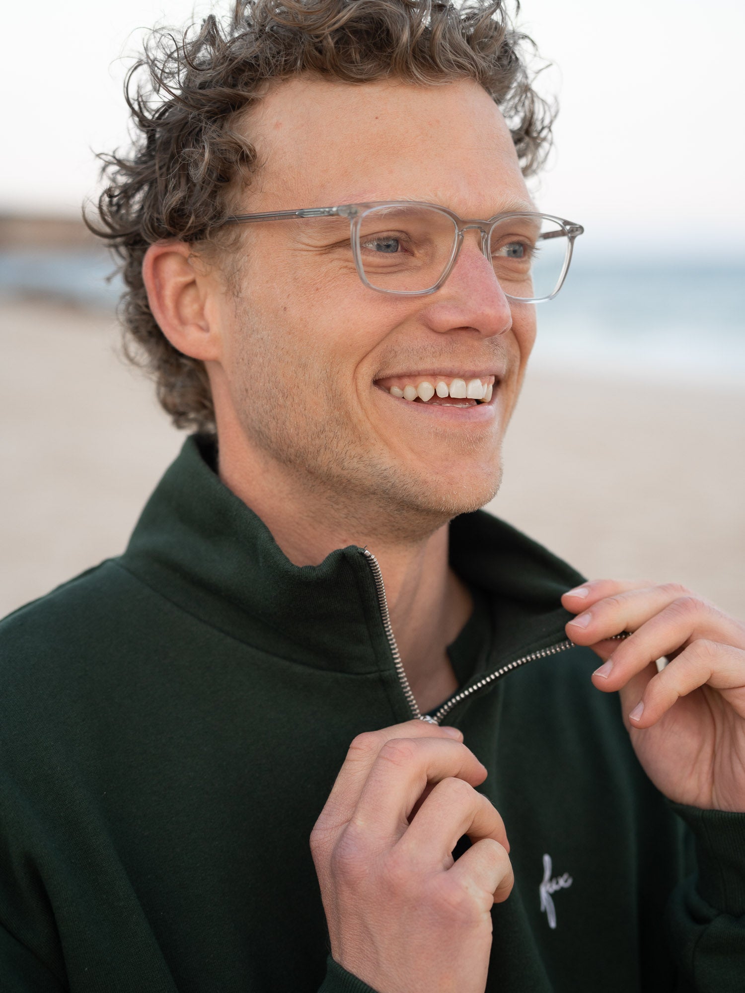 Junger, blonder Mann trägt einen Fair Fashion Männer Half Zip Sweater von FUXBAU in grün aus gots zertifizierter Biobaumwolle Made in Portugal mit YKK Reisverschlüssen am Strand in Portugal.