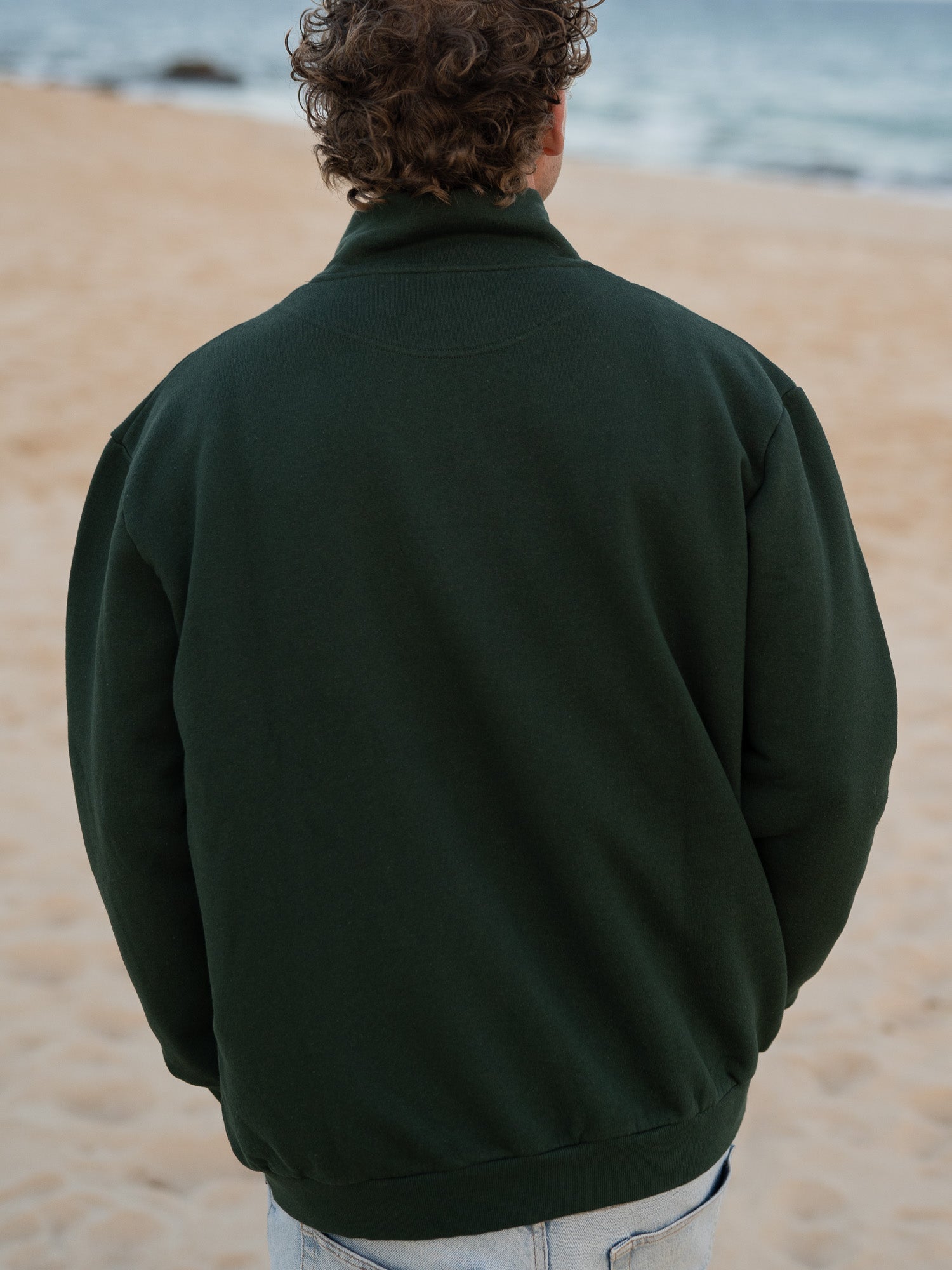 Junger, blonder Mann trägt einen Fair Fashion Männer Half Zip Sweater von FUXBAU in grün aus gots zertifizierter Biobaumwolle Made in Portugal mit YKK Reisverschlüssen am Strand in Portugal.