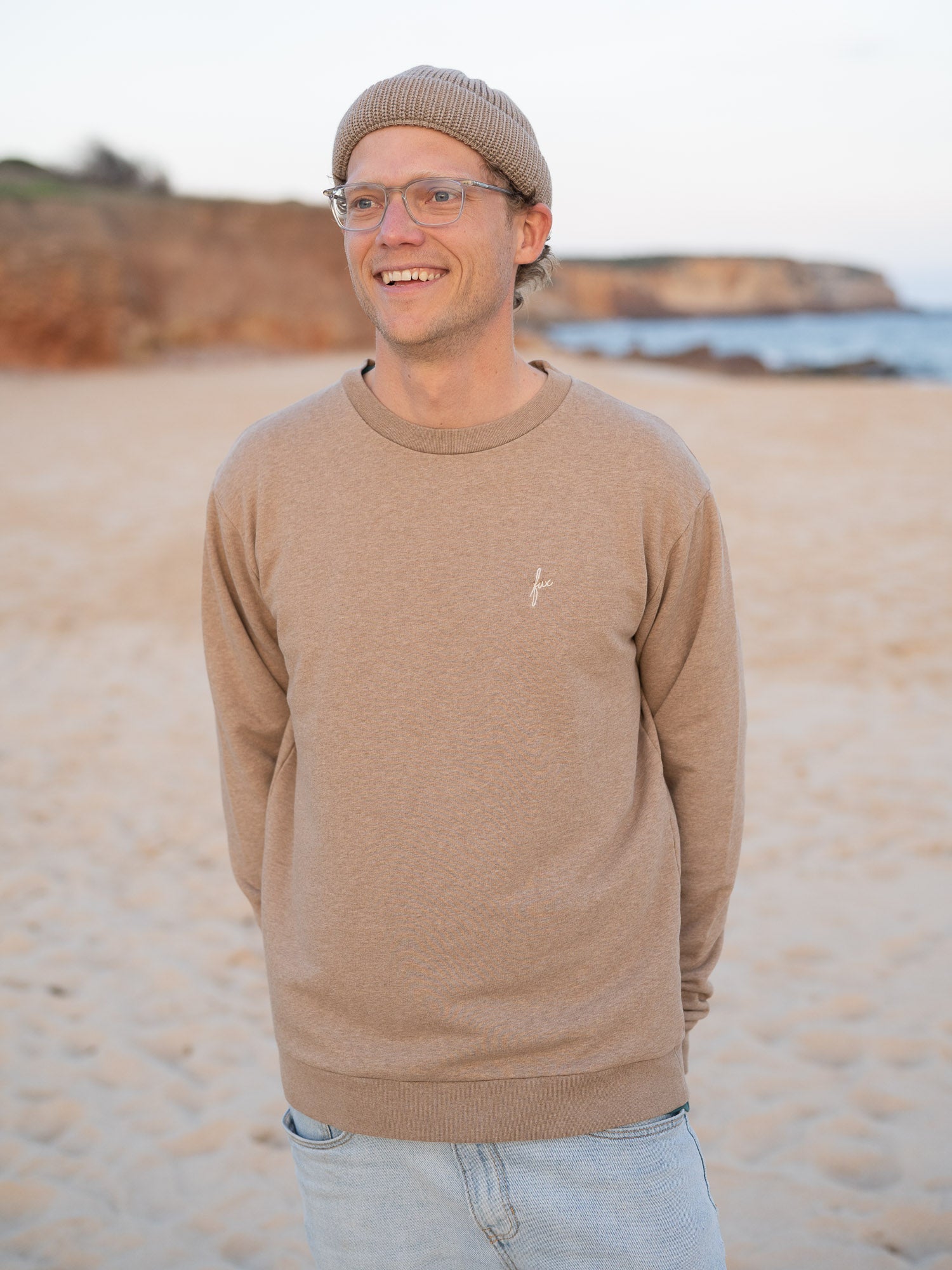 Nachhaltiger Fair Fashion Männer fux Sweater in beige meliert von FUXBAU aus 100% Biobaumwolle getragen am Strand in Portugal.