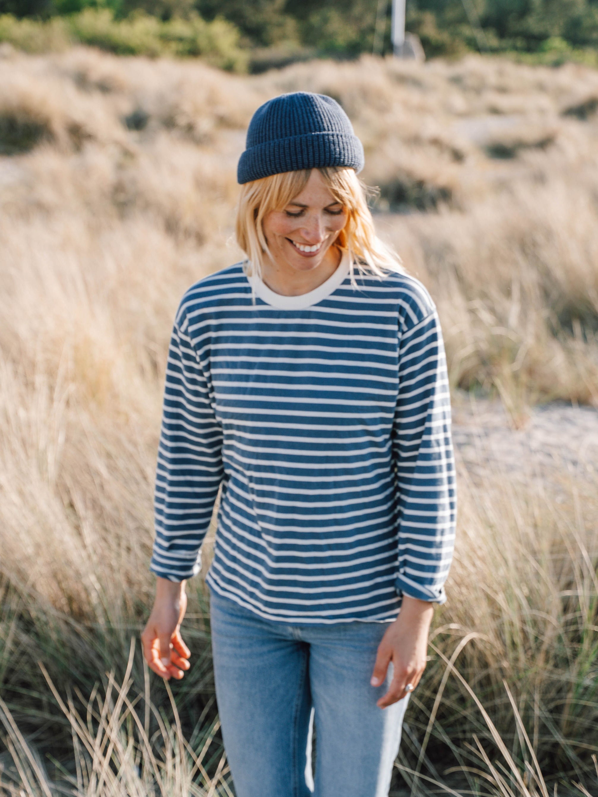 Junge, blonde Frau trägt ein maritimes FUXBAU Fair Fashion Outfit am Strand bestehend aus einem gestreiftem Longsleeve in creme blau, jeans und fischermütze aus Bio-Baumwolle.