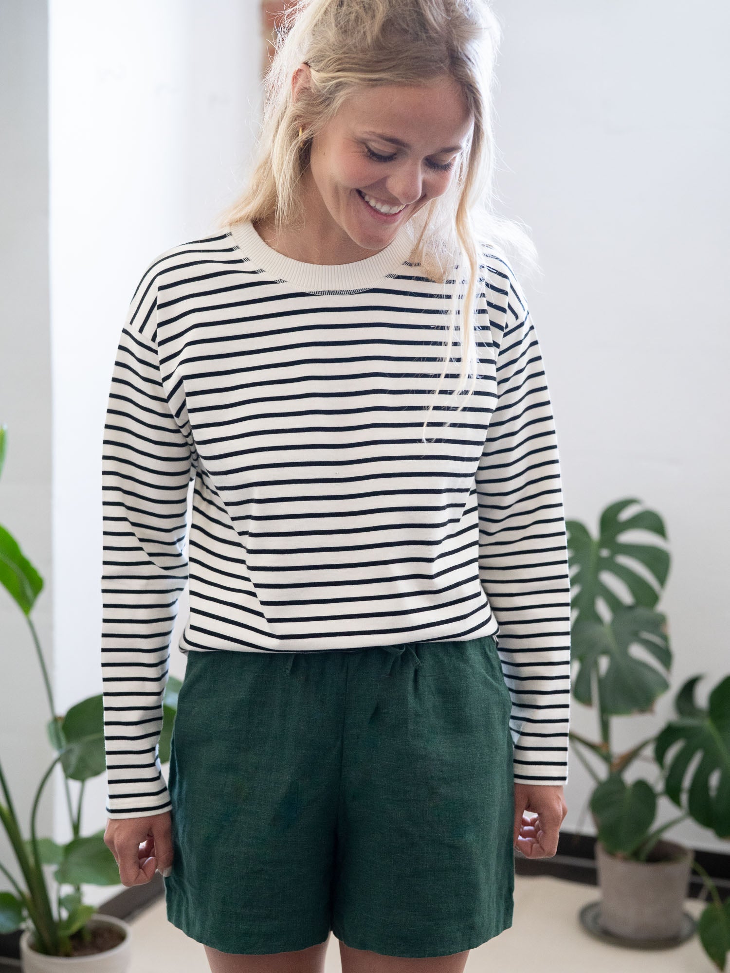 Junge blonde Frau trägt ein Fair Fashion Outfit von FUXBAU, bestehend aus einer grünen Leinenshorts und einem weiß schwarz gestreiftem Longsleeve.