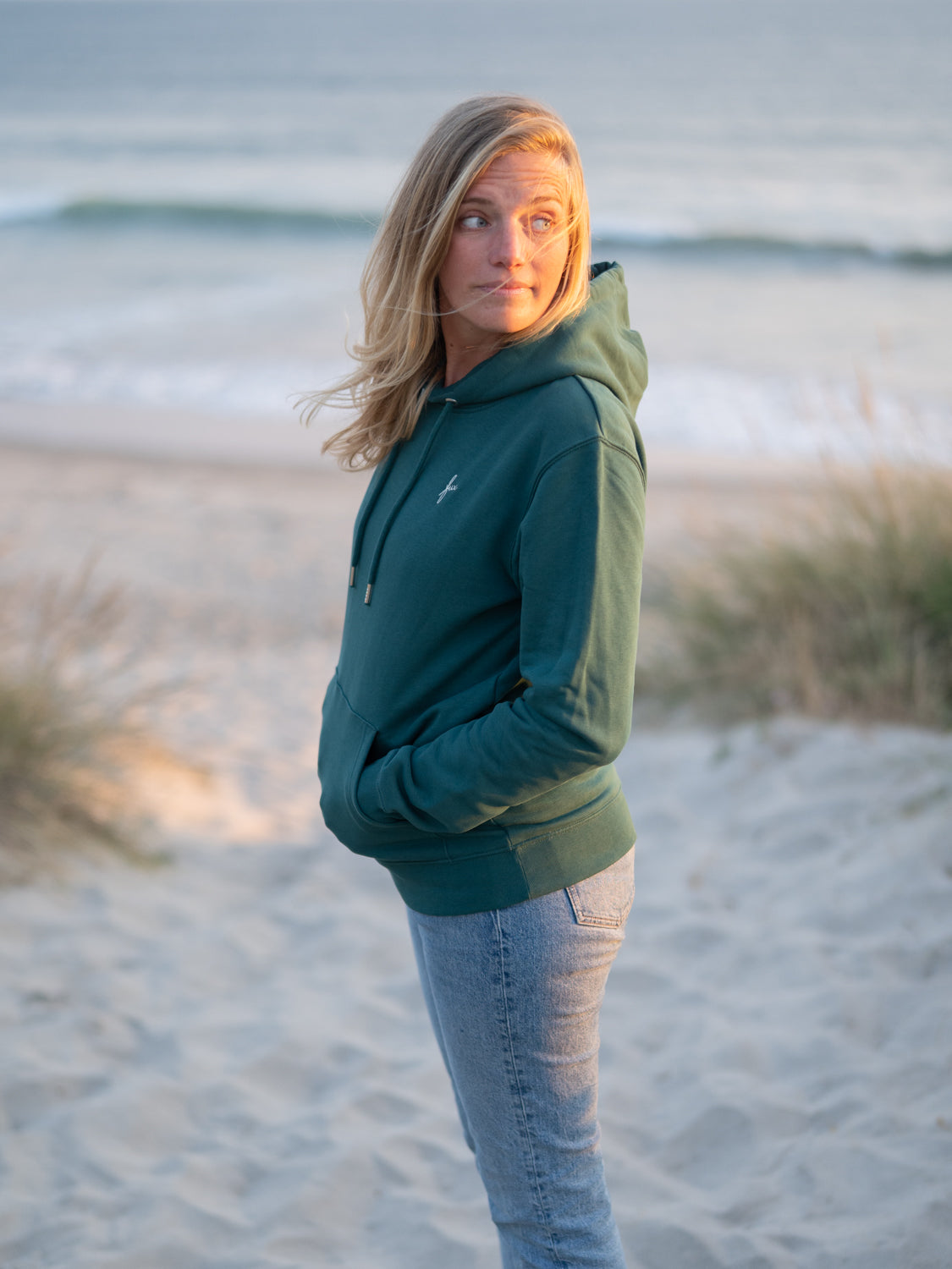 Eine junge, blonde Frau trägt einen fair produzierten grünen FUXBAU Unisex Hoodie aus Biobaumwolle am Strand in Portugal.