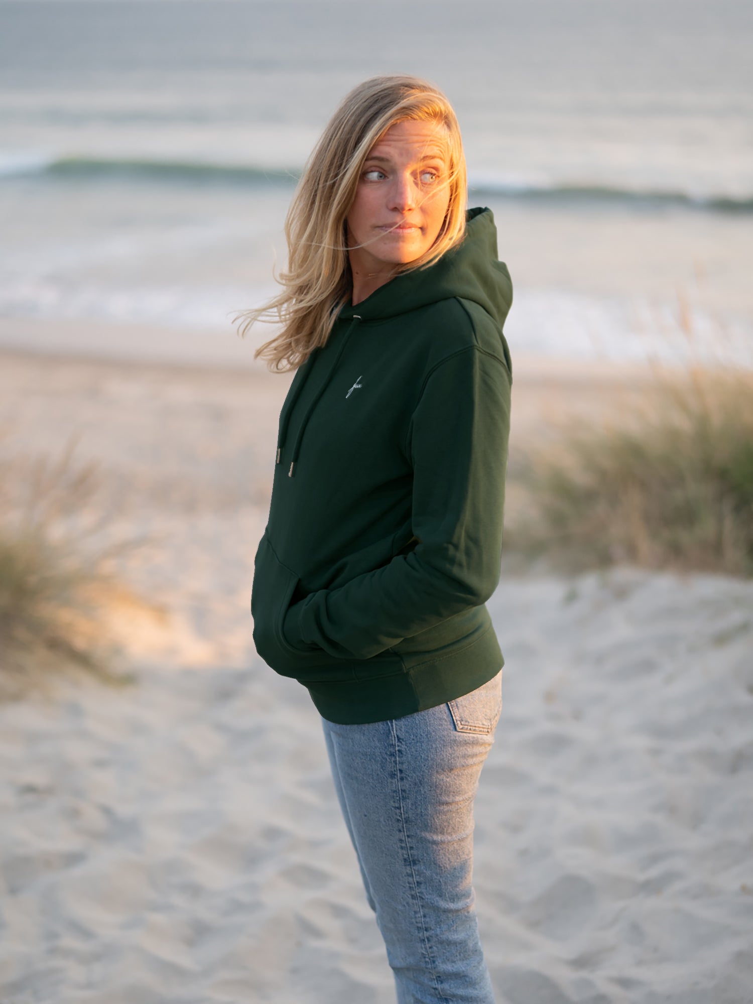 Eine junge, blonde Frau trägt einen fair produzierten grünen FUXBAU Unisex Hoodie aus Biobaumwolle am Strand in Portugal.
