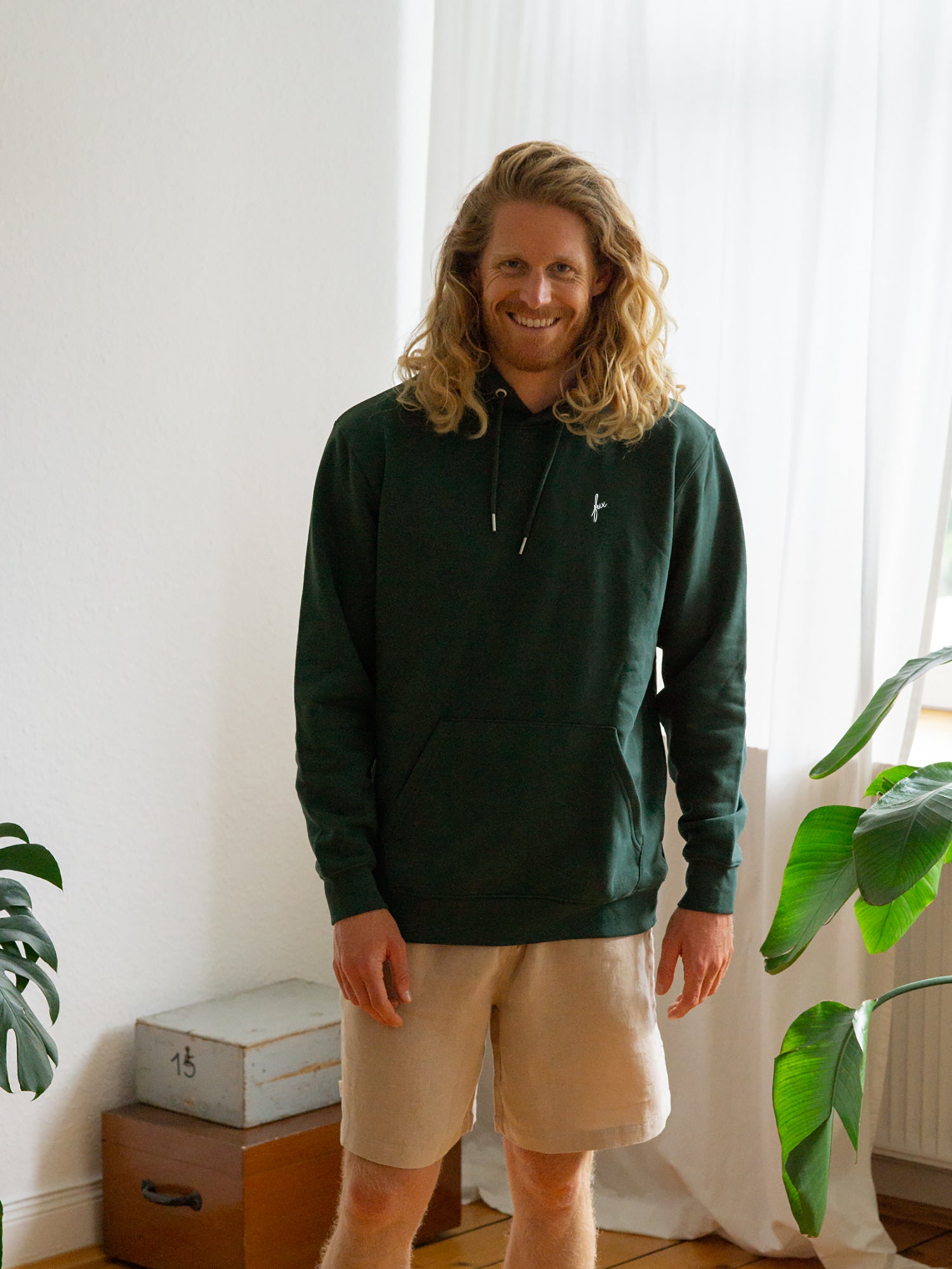 Junger, langhaariger blonder Mann trägt einen fair produzierten Hoodie in grün von FUXBAU