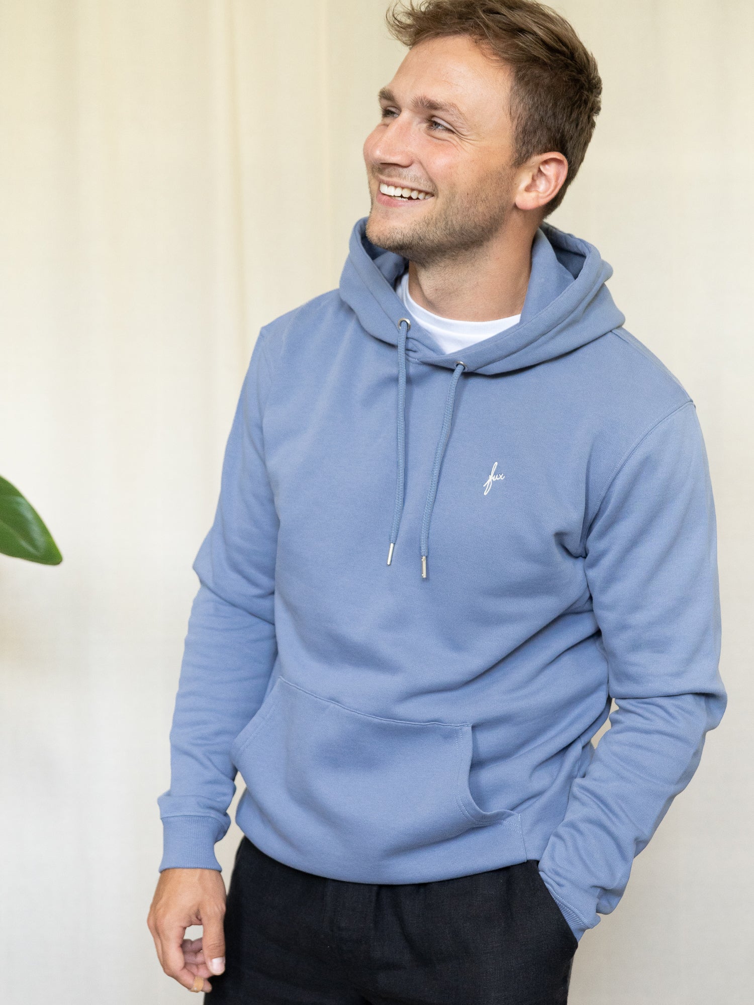 Frontalansicht eines Mannes mit himmelblauem Hoodie aus Biobaumwolle und schwarzer Hose, lächelnd.