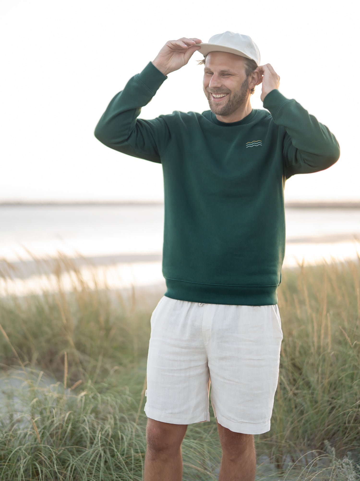 Nachhaltiger Fair Fashion Unisex Pullover von FUXBAU in grün mit einer Wellen Stickerei aus Biobaumwolle getragen von Philipp am Strand in Portugal.