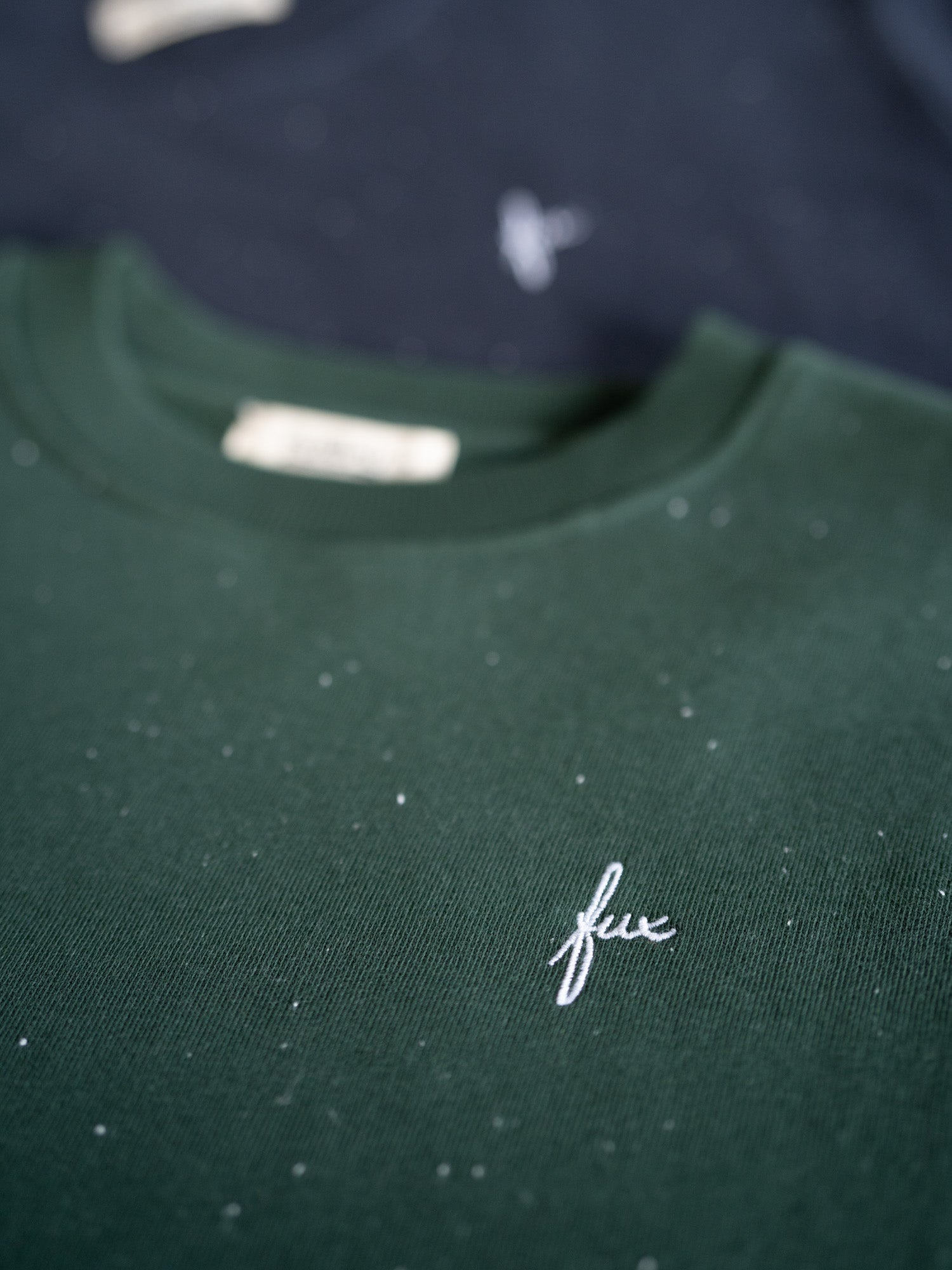 Grüner FUXBAU Neps Pullover aus nachhaltiger Bio-Baumwolle – gesticktes FUX Logo auf der Brust, im Hintergrund Variante in Navy