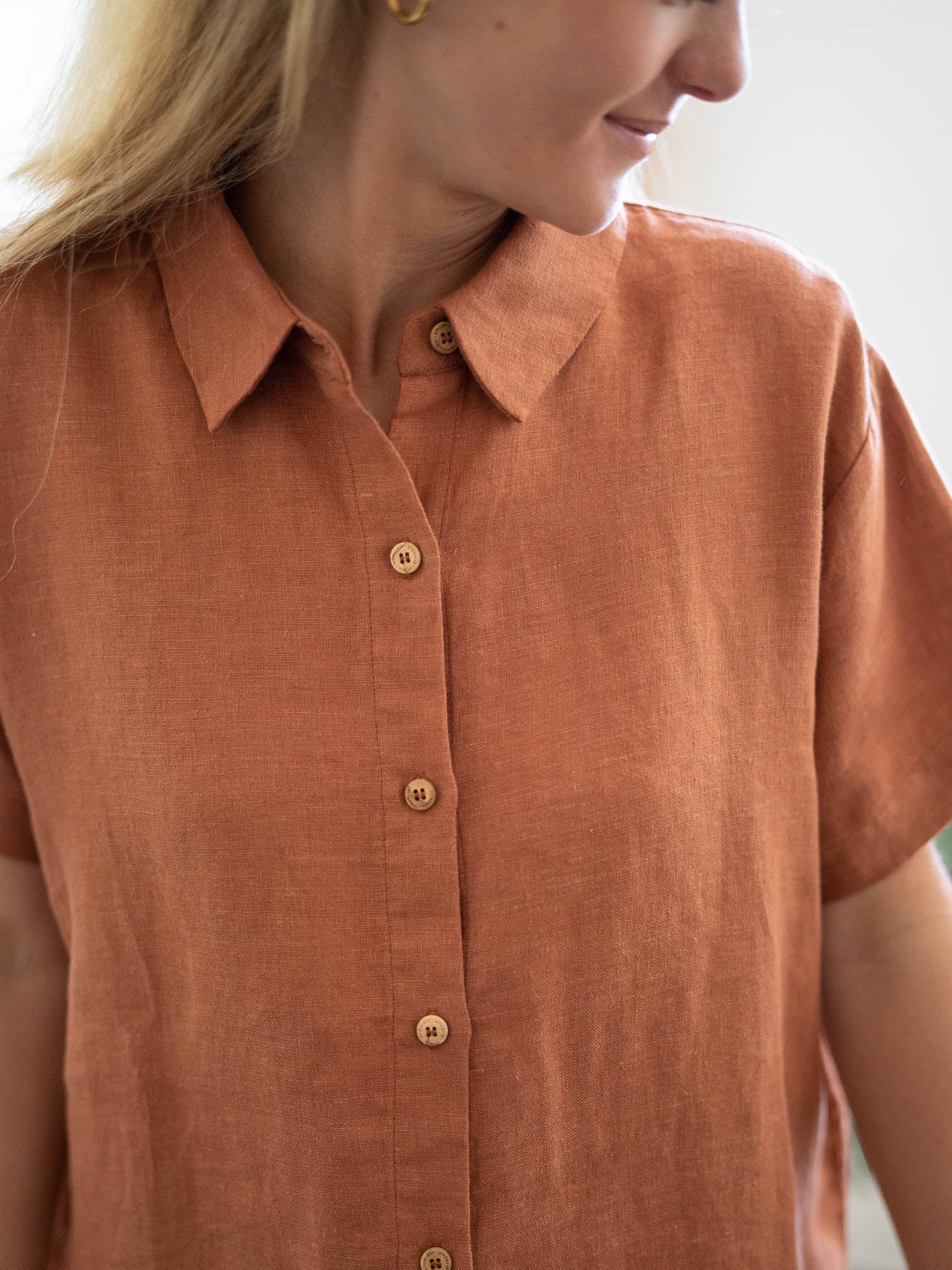 Eine junge, blonde Frau trägt eine nachhaltige Fair Fashion Kurzarm Leinenbluse in rotbraun von FUXBAU. Detailansicht der Knopfleiste.
