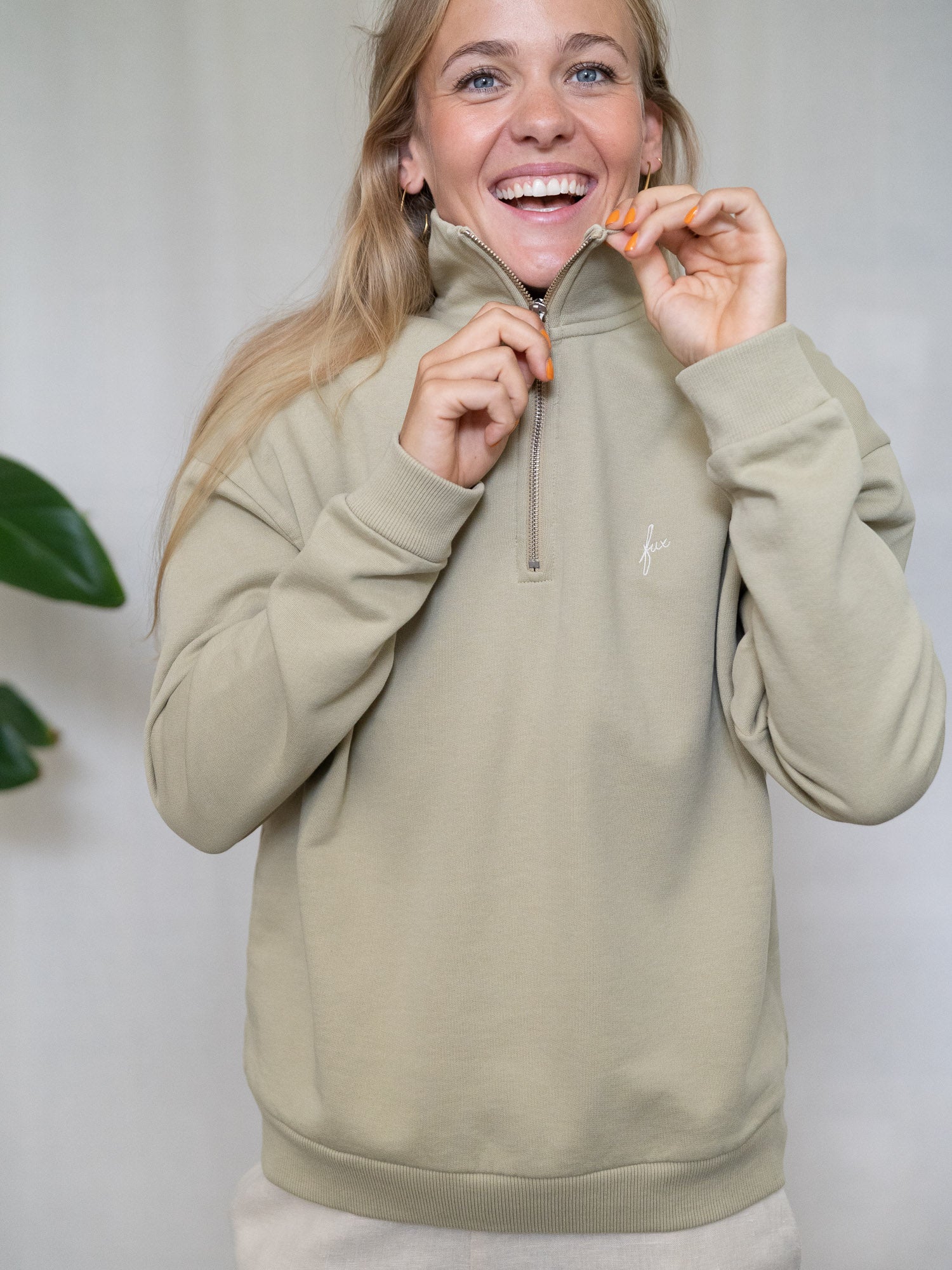Person öffnet lächelnd den Reißverschluss eines salbeifarbenen Half-Zip-Sweaters aus Biobaumwolle