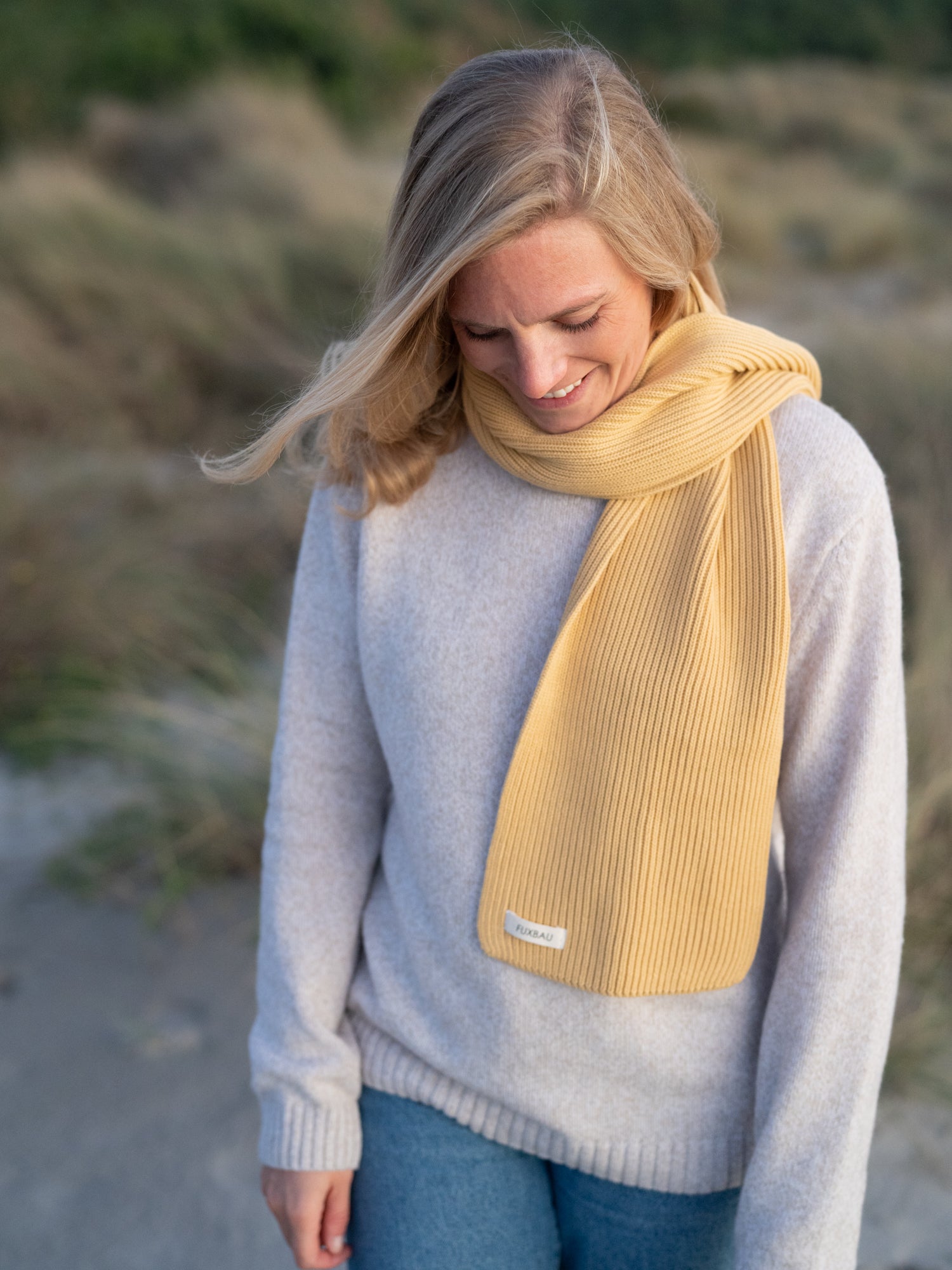 Junge, blonde Frau trägt einen gelben fair fashion Strickschal von FUXBAU aus GOTS zertifizierter Biobaumwolle am Strand in Portugal.