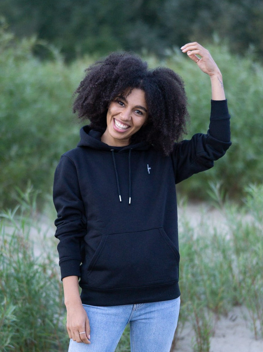 Junge, schwarzhaarige Frau trägt einen fairen und nachhaltigen Hoodie von FUXBAU in schwarz in Hamburg am Strand.
