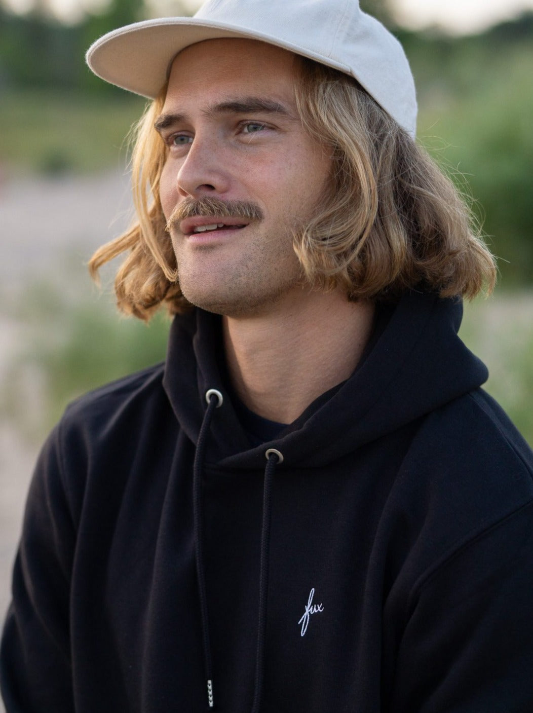 Junger, braunhaariger Mann trägt einen schwarzen FUXBAU Hoodie aus Biobaumwolle.