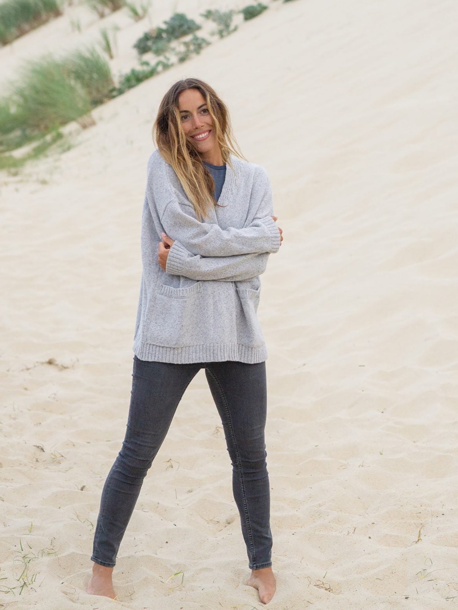 Graumelierter Oversized Frauen Cardigan aus 100% Merino Schurwolle von FUXBAU. Getragen von einem braunhaarigem Model am Strand in Fankreich.