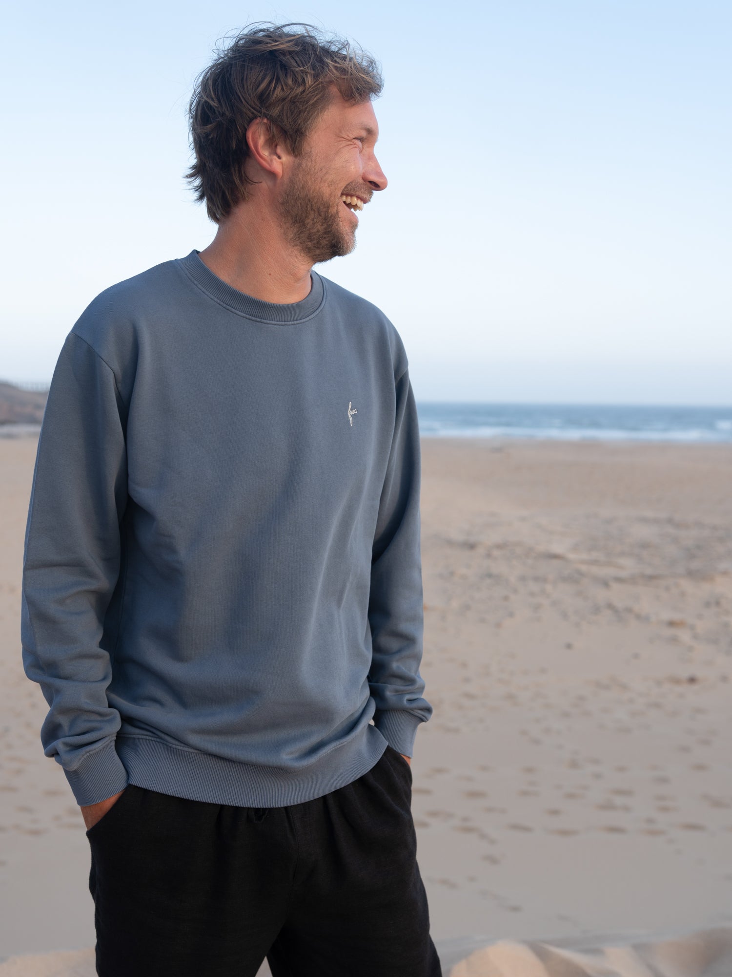 Mann trägt fair gefertigten seeblauen FUXBAU Pullover aus Biobaumwolle vor Strand und Meer
