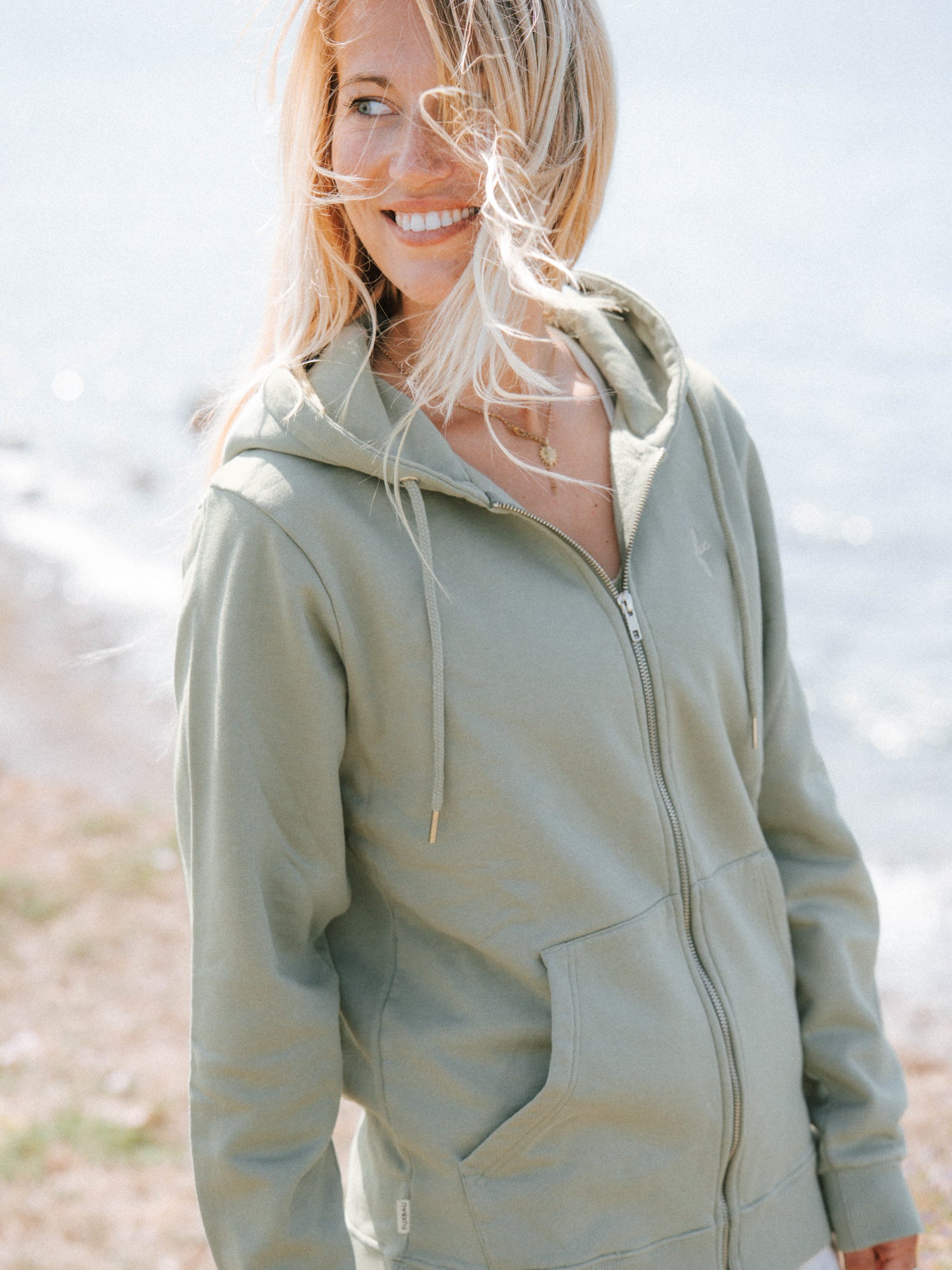 Unisex Sweatjacke in Seegras aus Biobaumwolle in seitlicher Ansicht am Meer
