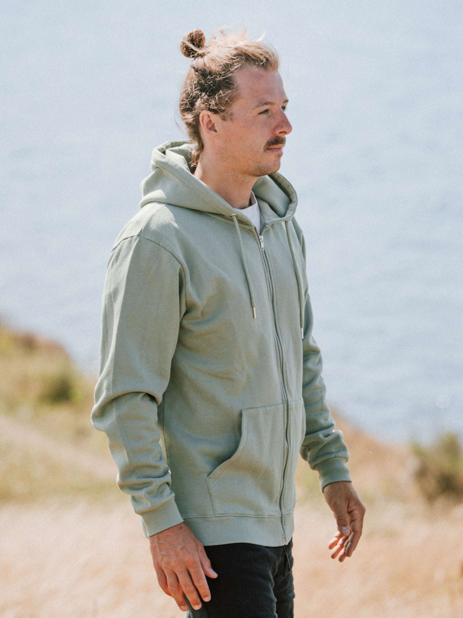 Unisex Sweatjacke in Seegras aus Biobaumwolle getragen von einem Mann an der Küste