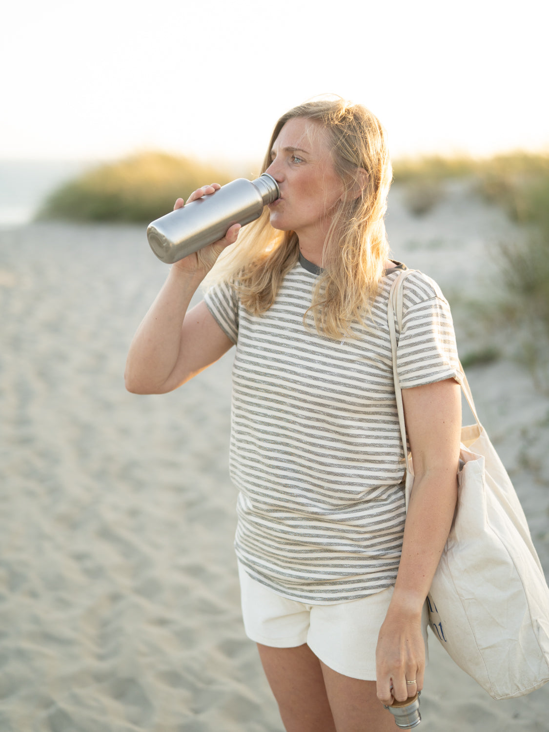 Junge, blonde Frau steht am Strand in Portugal mit einem Fair Fashion Outfit und einer nachhaltigen Trinkflasche aus Edelstahl und Bambusdeckel von FUXBAU.