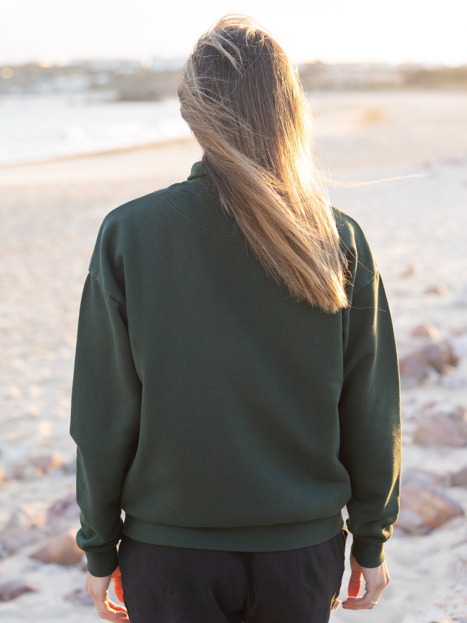 Junge, braunhaarige Frau trägt einen Fair Fashion Frauen Half Zip Sweater von FUXBAU in grün aus gots zertifizierter Biobaumwolle Made in Portugal mit YKK Reisverschlüssen am Strand in Portugal.