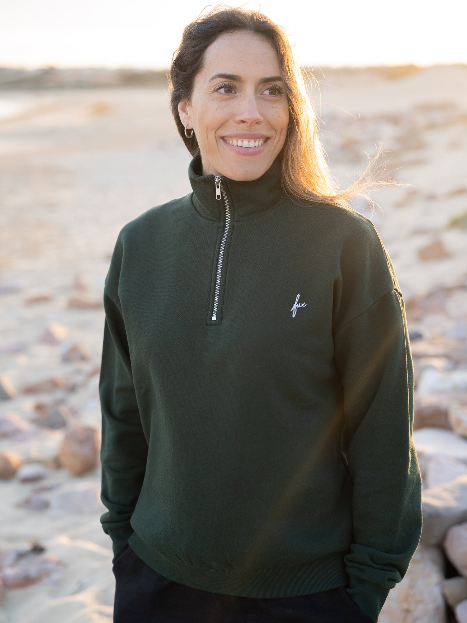 Junge, braunhaarige Frau trägt einen Fair Fashion Frauen Half Zip Sweater von FUXBAU in grün aus gots zertifizierter Biobaumwolle Made in Portugal mit YKK Reisverschlüssen am Strand in Portugal.