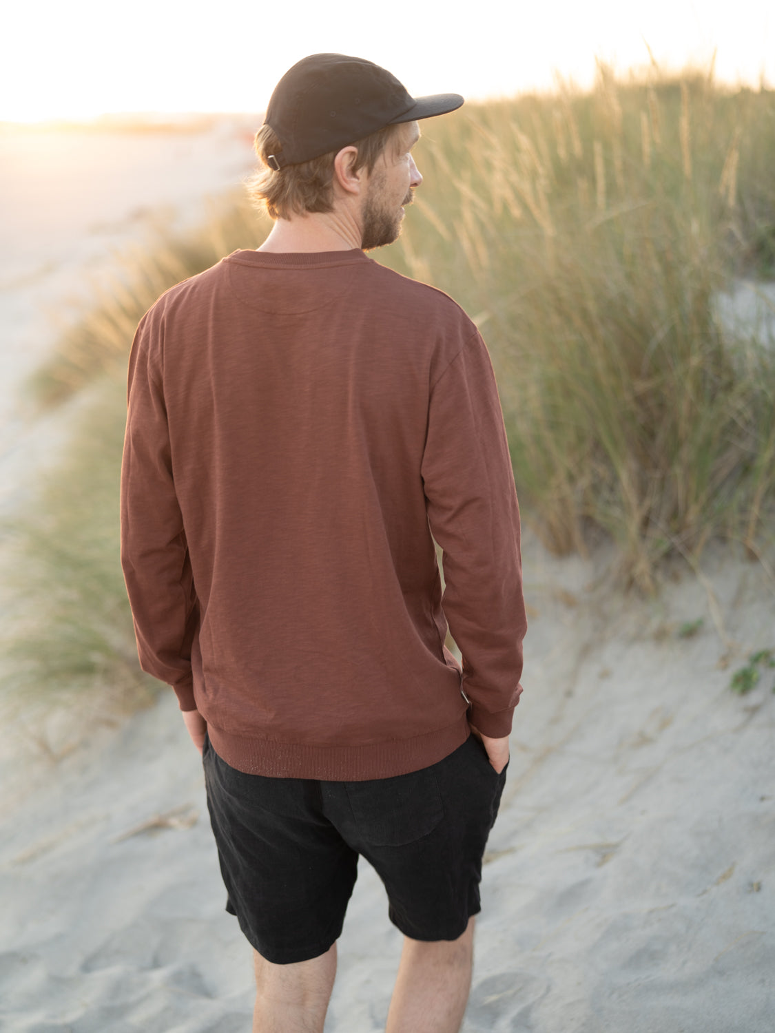 Kenny trägt den nachhaltigen und fair produzierten Männer fux Sweater in braun aus 100% GOTS zertifizierter Biobaumwolle am Strand.