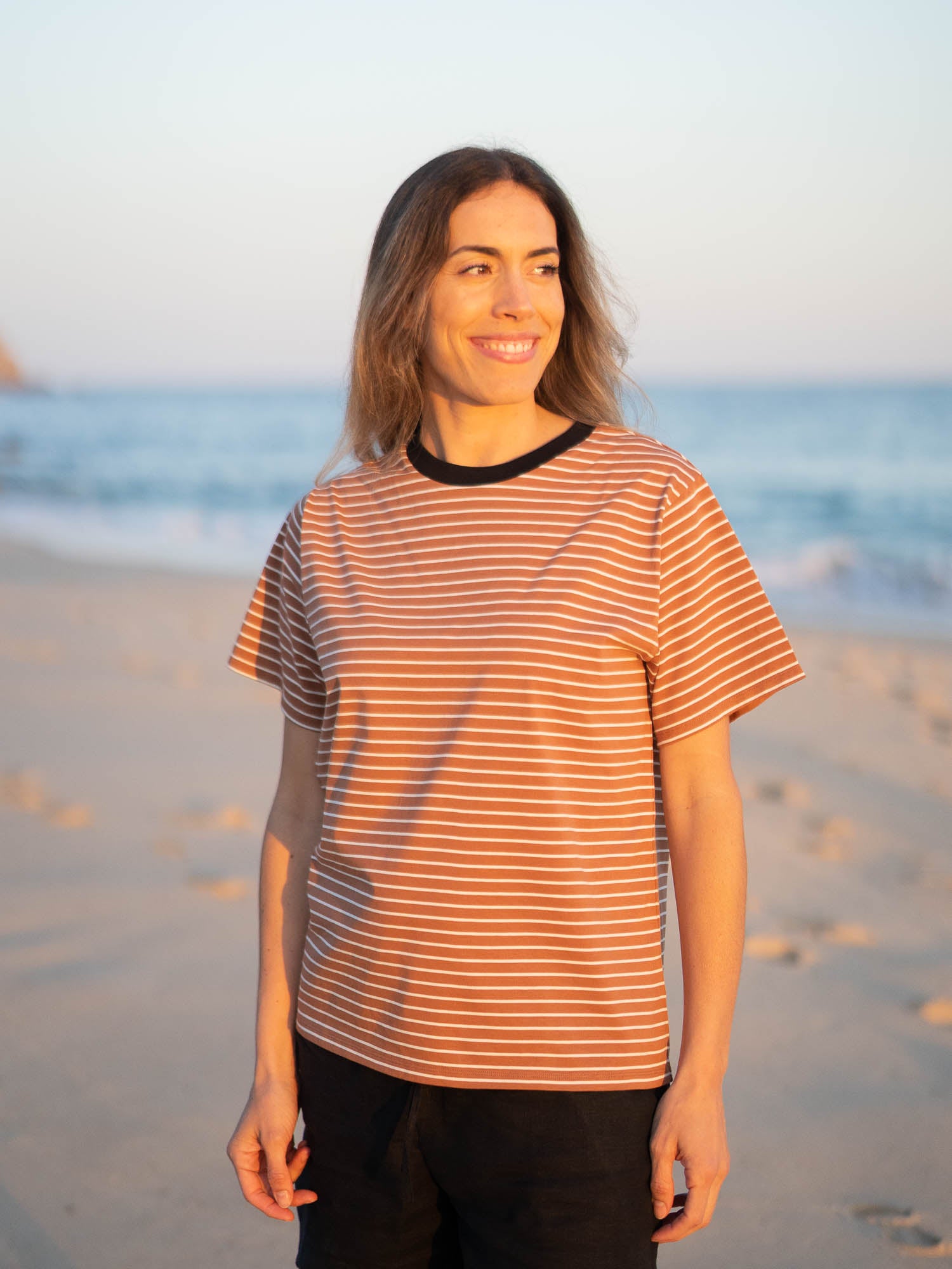 Gestreiftes Fair Fashion Frauen T-Shirt in karamell, schwarz und weiss von FUXBAU aus Biobaumwolle. Gefertigt unter nachhaltigen GOTS Bedingungen in Portugal und getragen am Strand in Portugal. Getragen von einer jungen Frau am Strand in Südportugal.