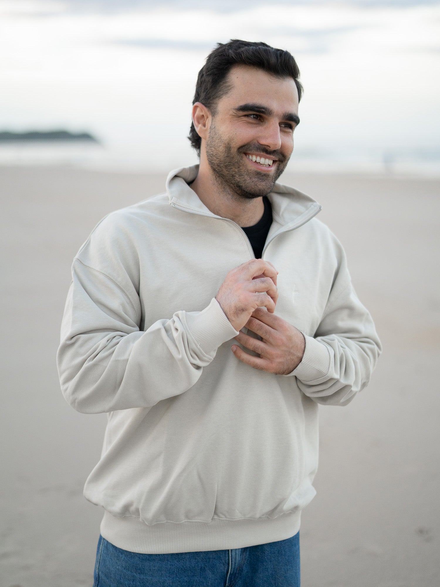 Ein nachhaltiger Fair Fashion Pullover mit Reißverschluss in Beige von FUXBAU wird von einem jungen Mann mit schwarzen Haaren am Strand in Spanien getragen. 