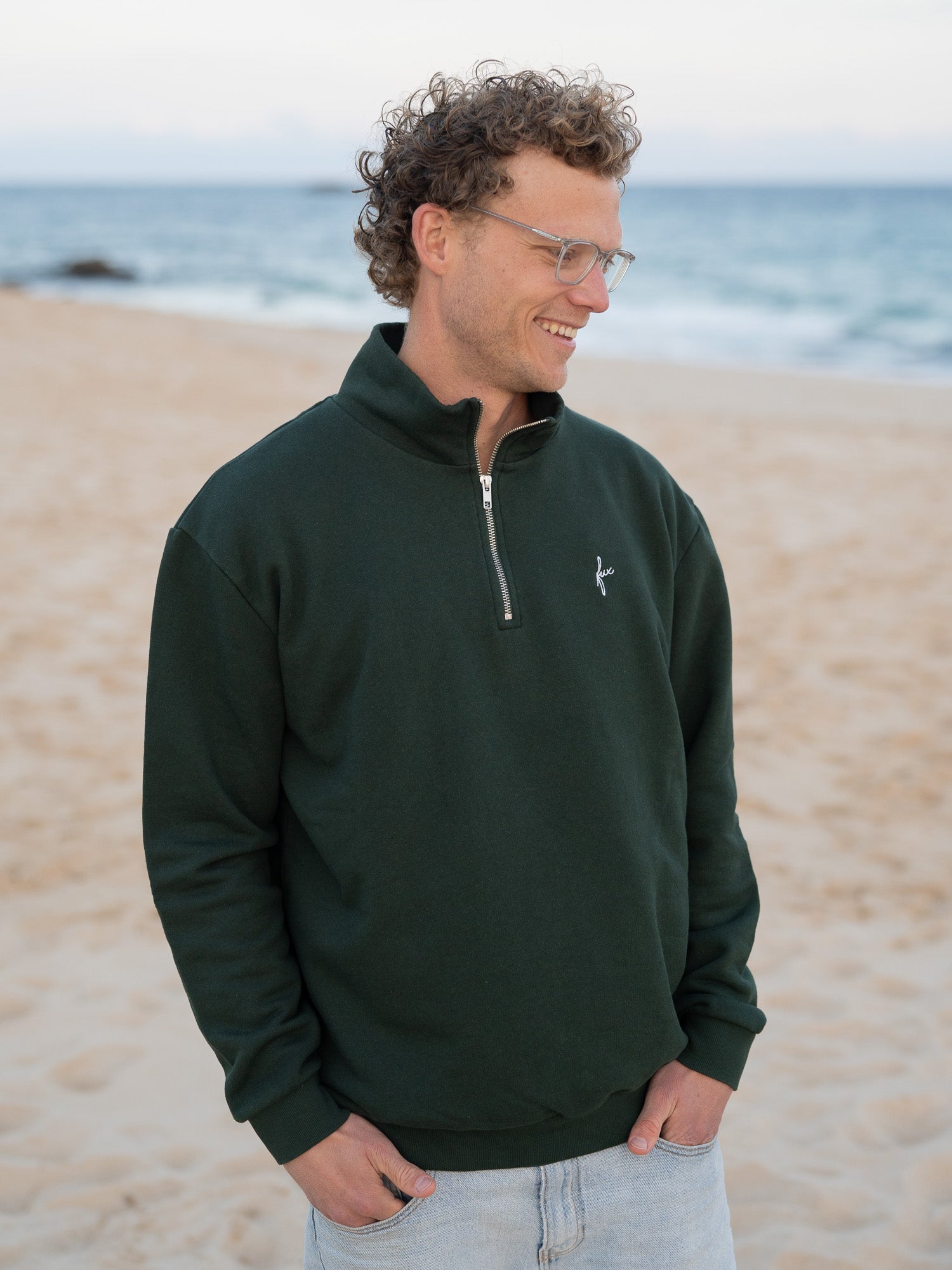 Junger, blonder Mann trägt einen Fair Fashion Männer Half Zip Sweater von FUXBAU in grün aus gots zertifizierter Biobaumwolle Made in Portugal mit YKK Reisverschlüssen am Strand in Portugal.