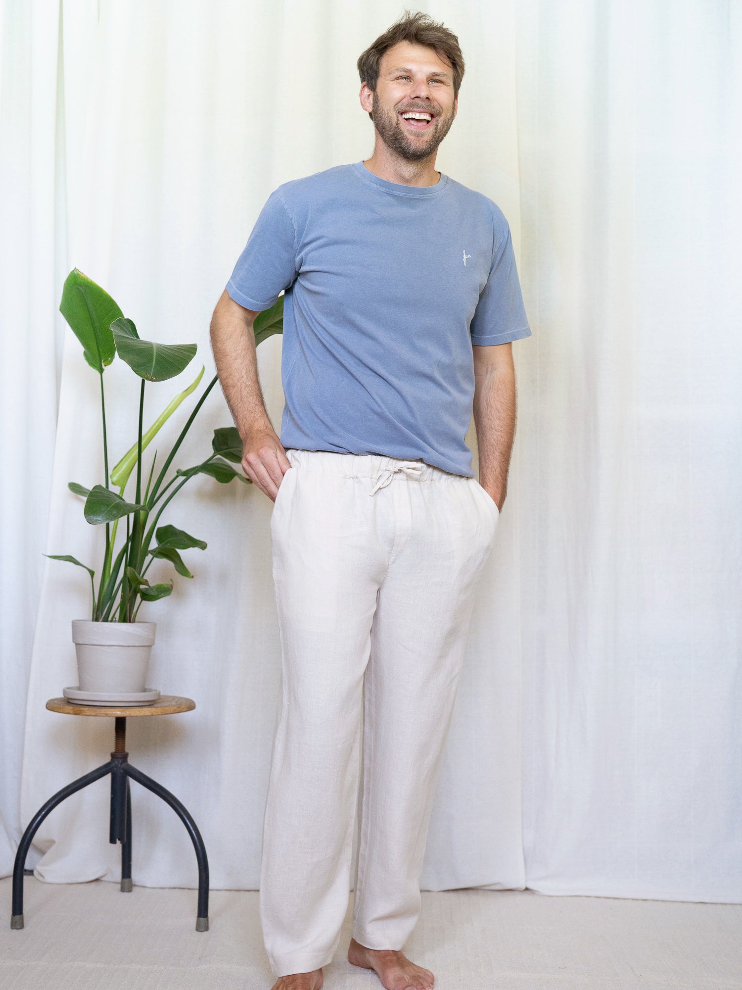 Ein junger Mann trägt eine nachhaltige Fair Fashion Leinenhose von FUXBAU in cremweiss in Kombination mit einem blauen T-Shirt aus Biobaumwolle vor einem Vorhang. 
