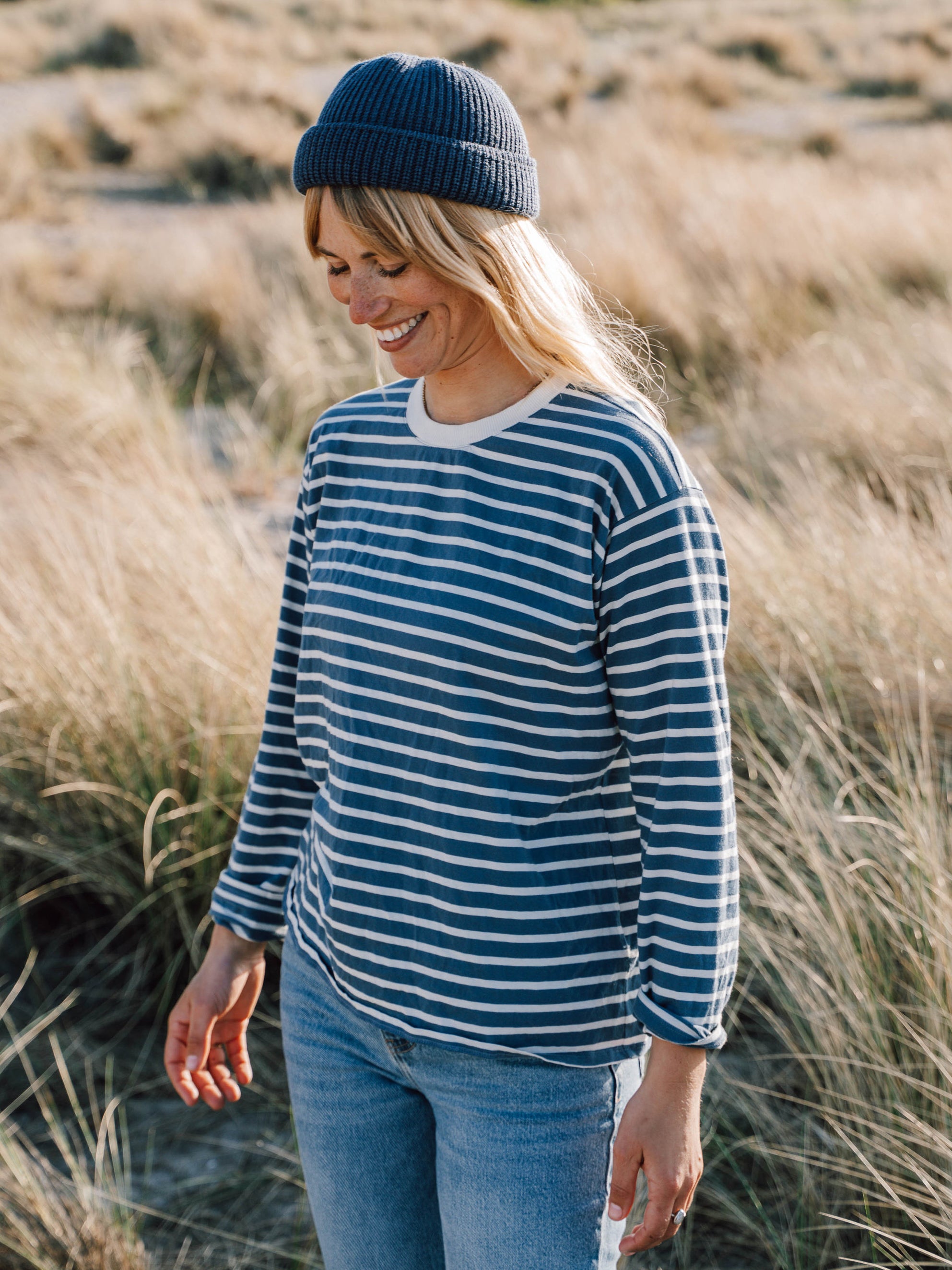 Junge, blonde Frau trägt ein maritimes FUXBAU Fair Fashion Outfit am Strand bestehend aus einem gestreiftem Longsleeve in creme blau, jeans und fischermütze aus Bio-Baumwolle.